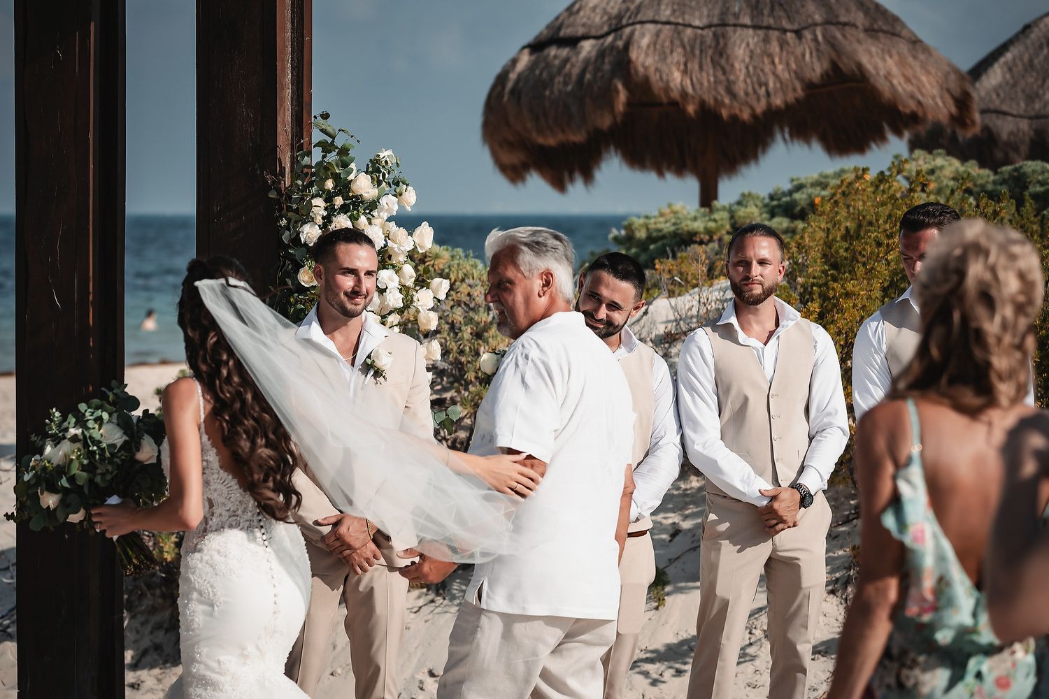 Beach wedding ceremony with bride, groom, and officiant near ocean, flanked by groomsmen and floral arch under thatched umbrella.