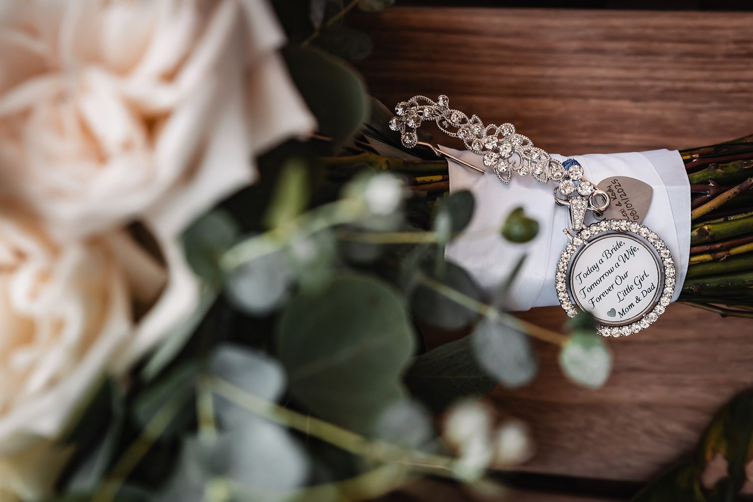 Bridal bouquet with white roses, eucalyptus, and a rhinestone memorial charm pendant on wood.