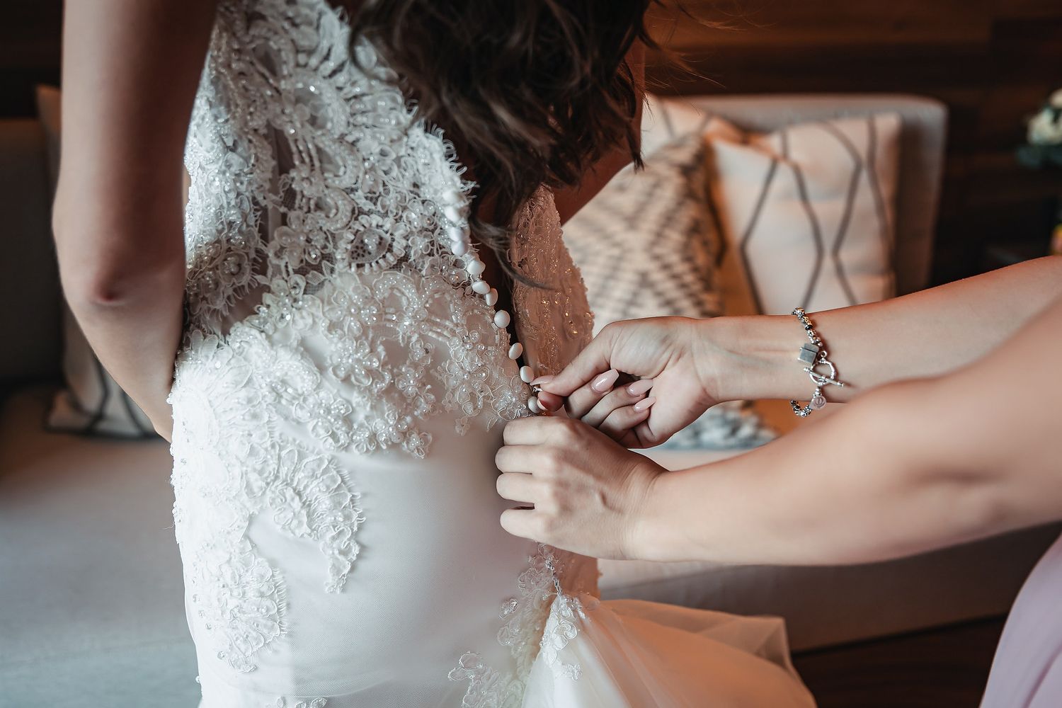 Bridesmaid fastening buttons on back of white lace wedding dress during bridal preparation.