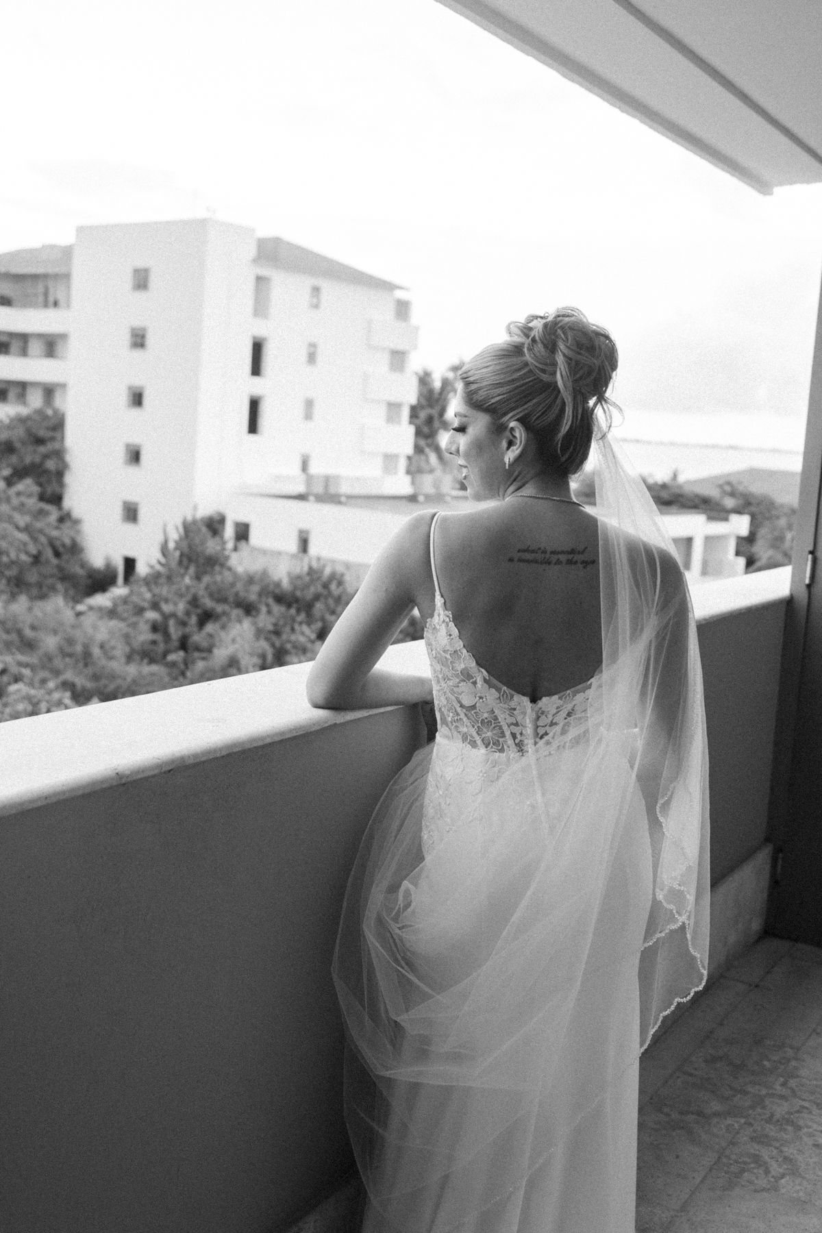 Bride in lace wedding dress and veil stands on balcony overlooking buildings, back tattoo visible, black and white photo.