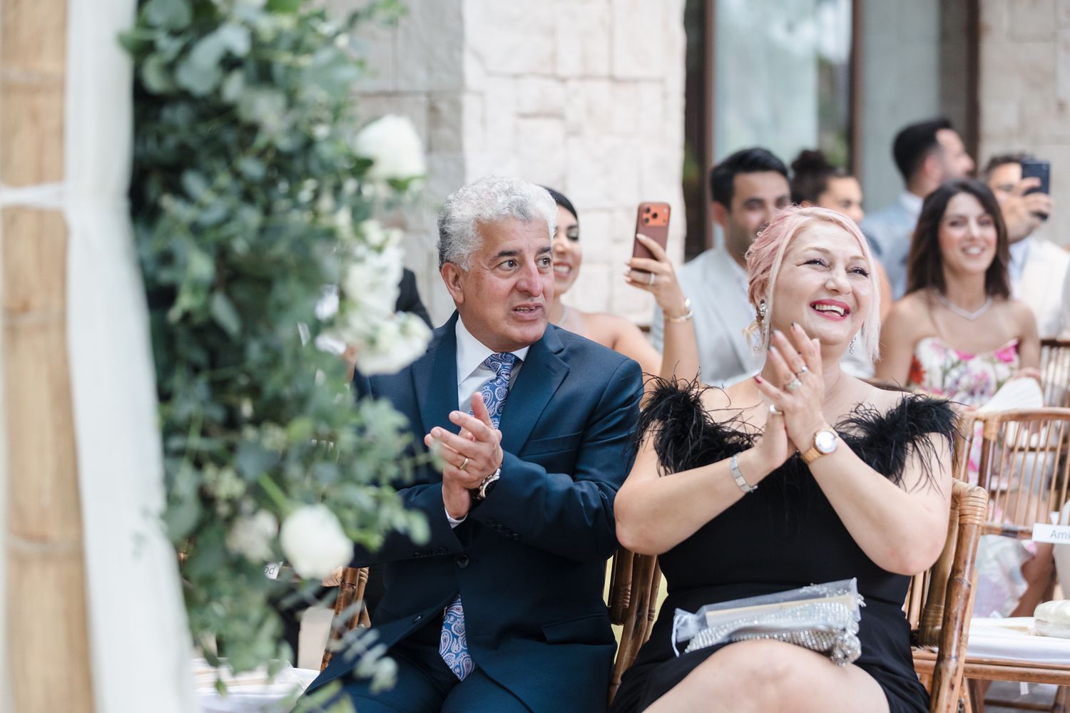 Guests clapping and smiling at an outdoor wedding reception, man in blue suit and woman in black dress.