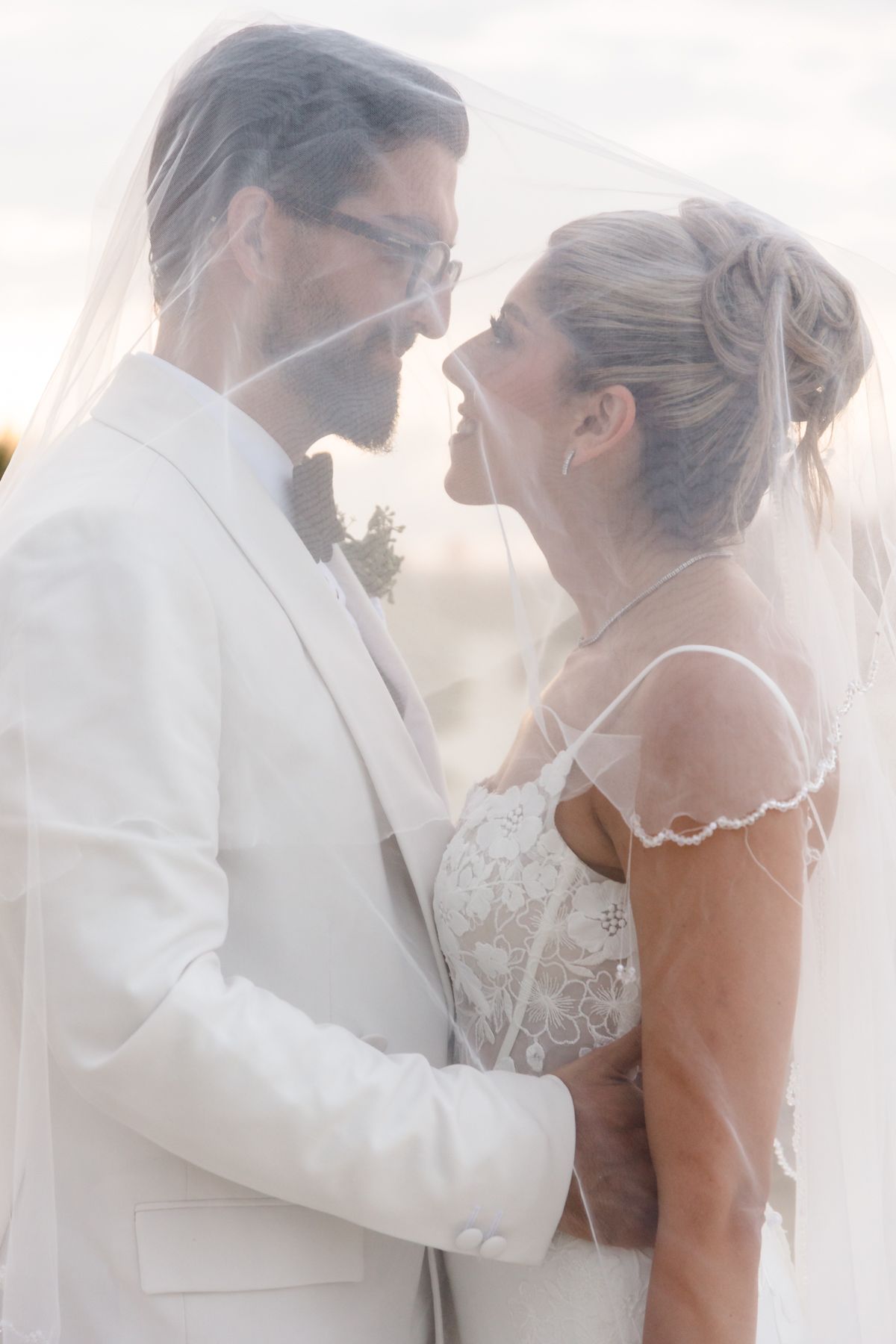 Bride and groom share a romantic moment under a sheer wedding veil, with the groom in a white suit and bride in lace gown.