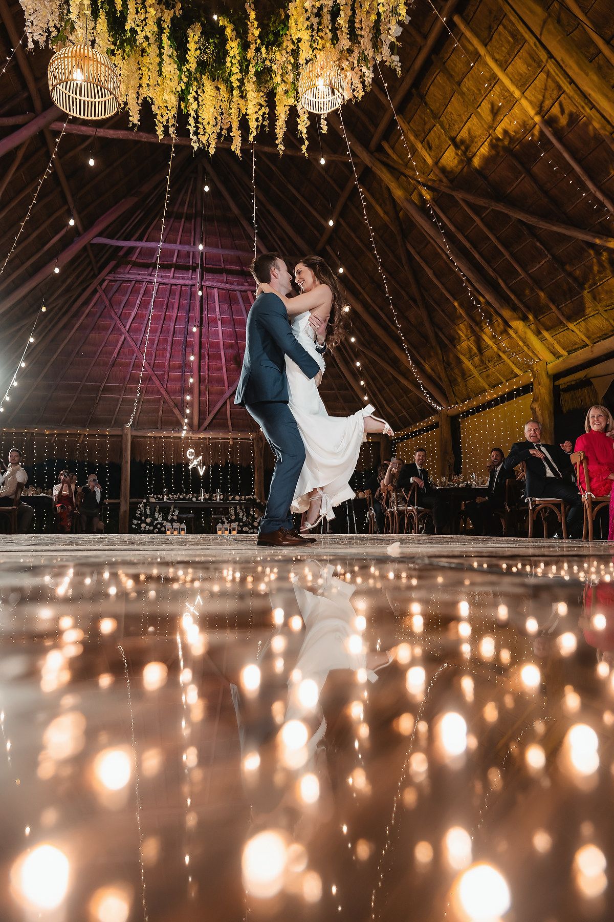 Groom lifting bride on candlelit dance floor in a rustic barn wedding venue with fairy lights and floral decorations.