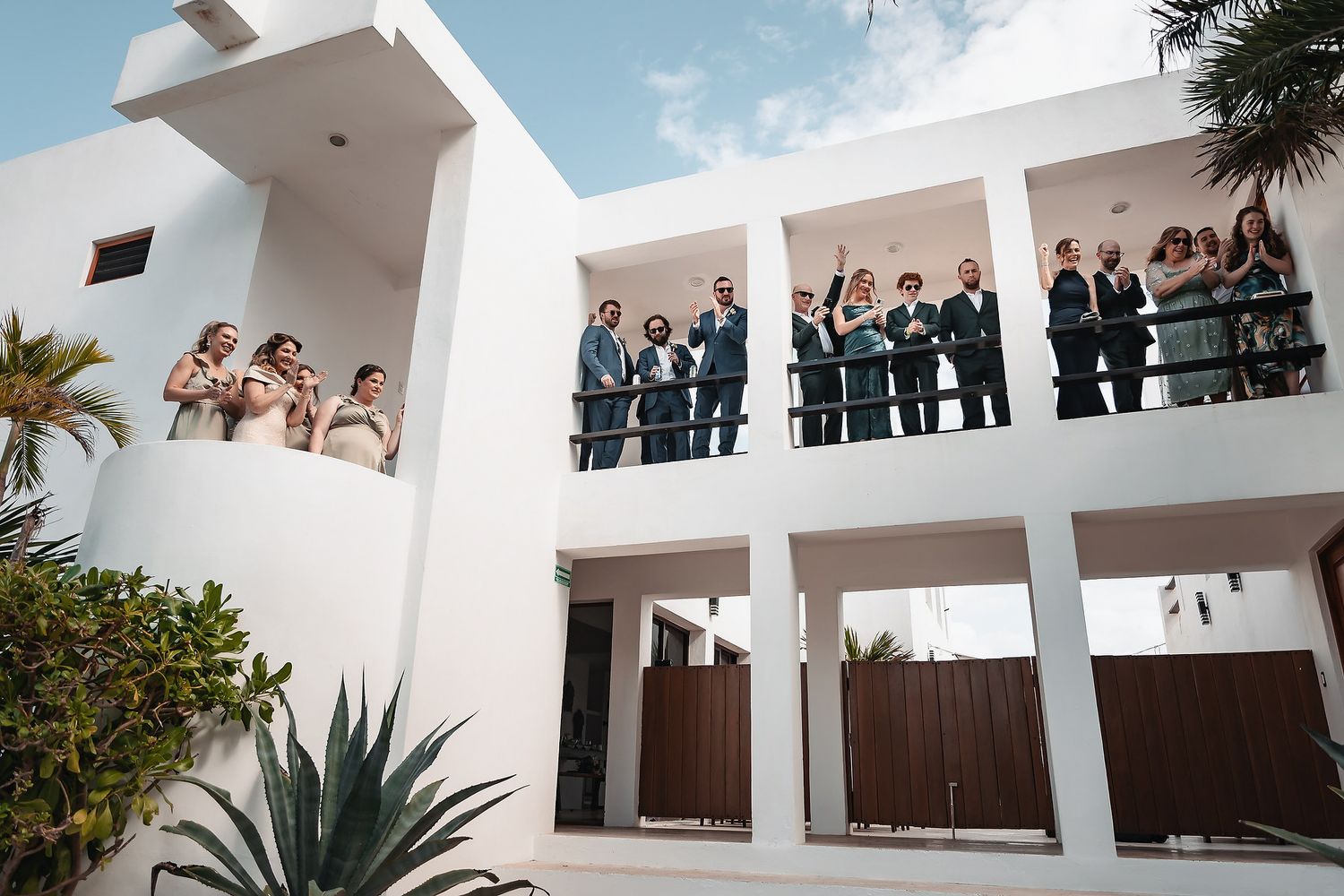 Wedding party gathered on white modern villa balcony, celebrating outdoors with agave plant in foreground.