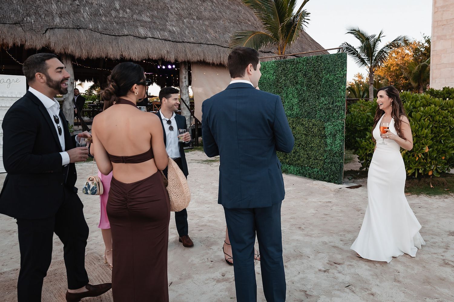 Wedding guests mingle on a sandy beach venue with a lush green hedge backdrop at a tropical destination wedding.