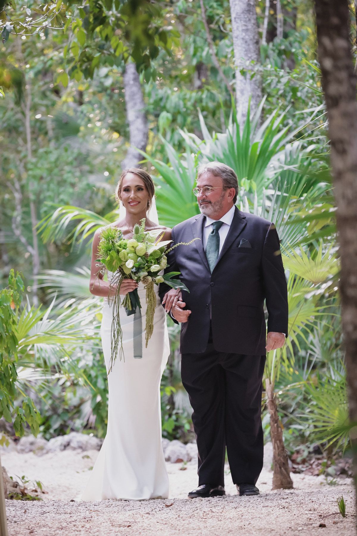 Bride in white gown holding green bouquet walks with older man in black suit through tropical garden path.