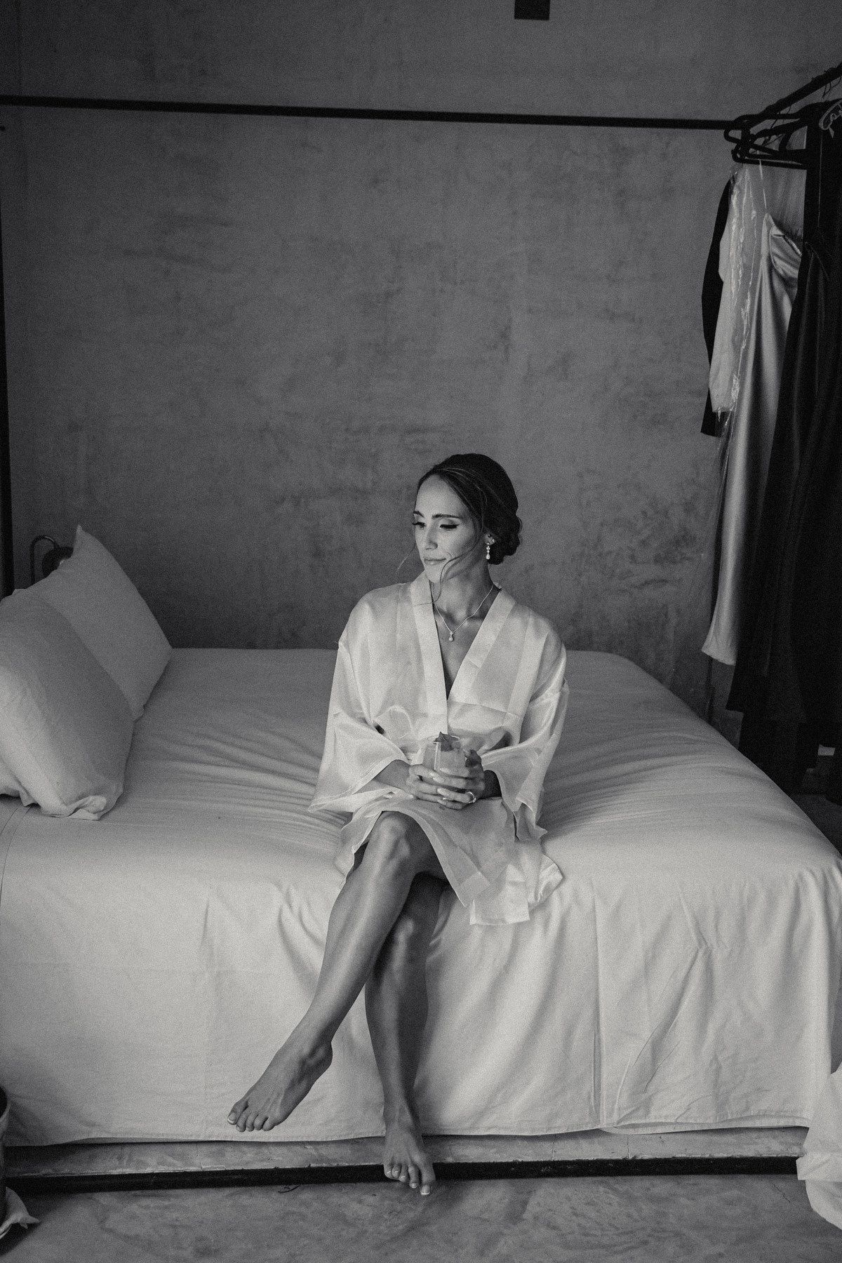 Elegant bride in white silk robe sits on bed holding a drink during pre-wedding preparations.