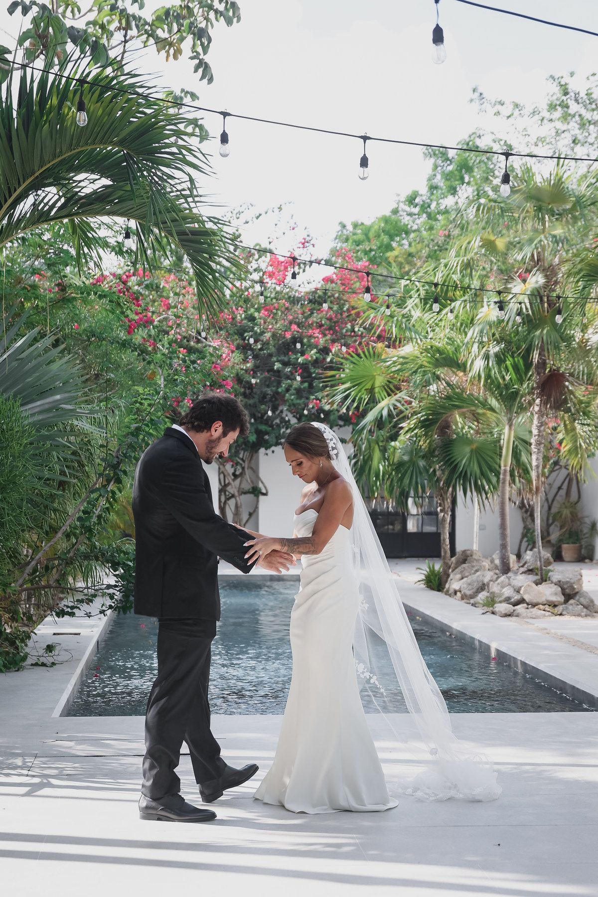 Bride and groom hold hands during first dance at tropical outdoor wedding venue with pool and lush greenery.
