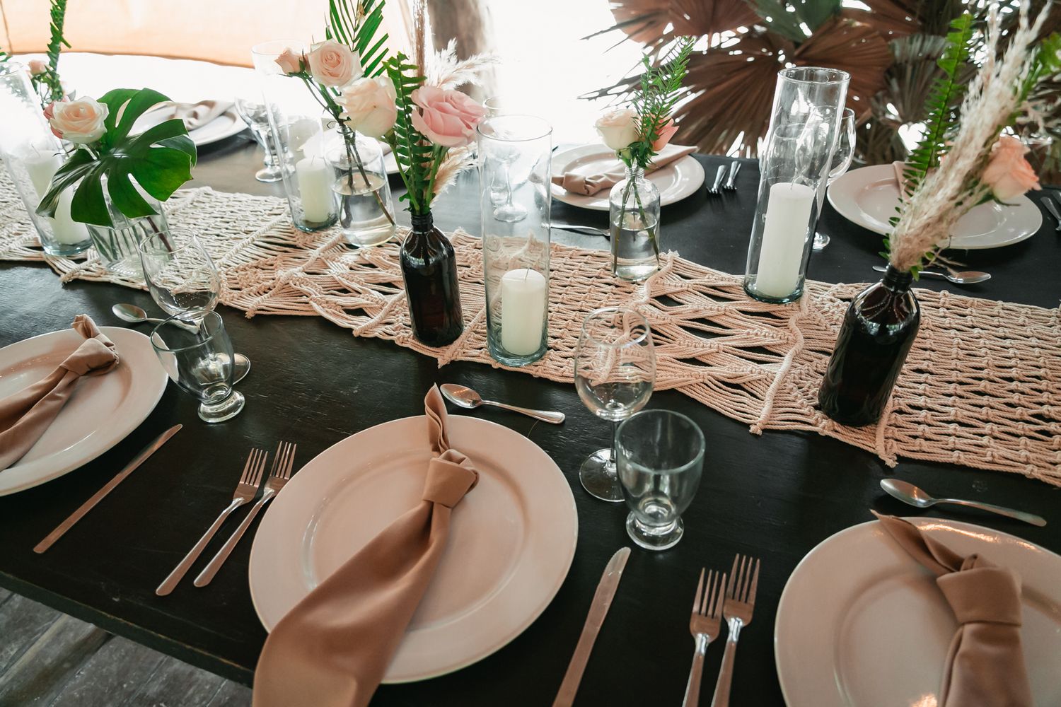 Tropical boho wedding reception table set with blush plates, macrame runner, candles, and floral centerpieces.