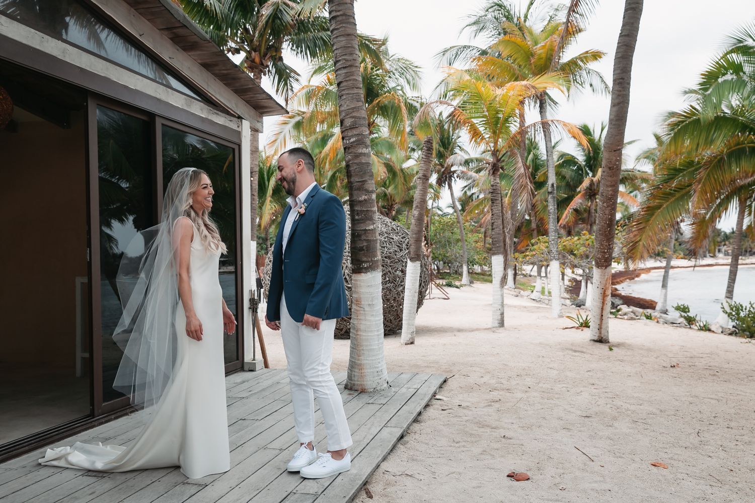 Bride in white gown and groom in blue suit hold hands on a beachside deck surrounded by lush tropical palm trees.