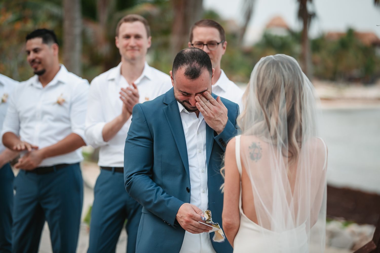 Emotional groom wiping tears during outdoor beach wedding ceremony with groomsmen standing behind him.