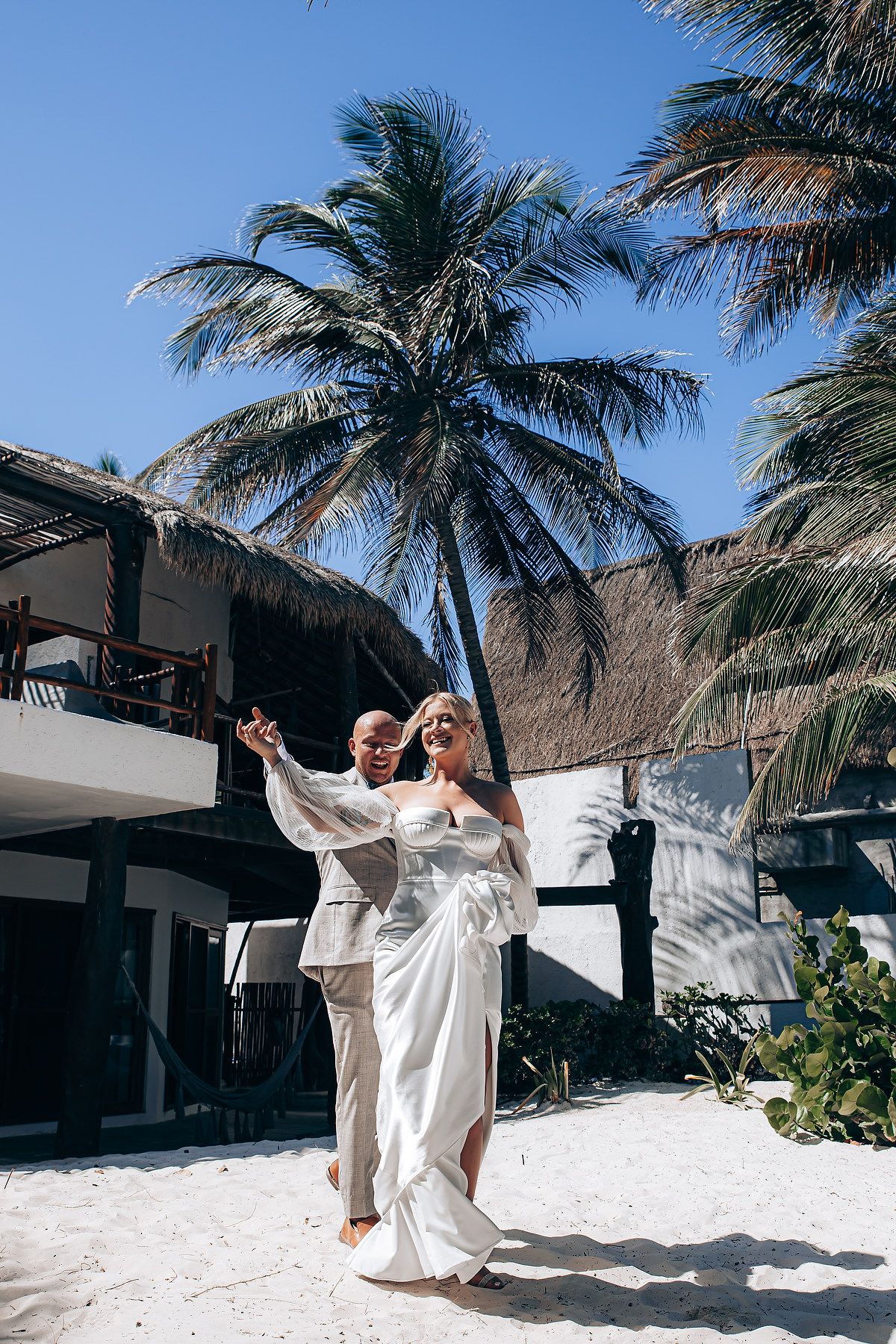 Beach wedding photoshoot with flowing white dresses against palm trees and sand.