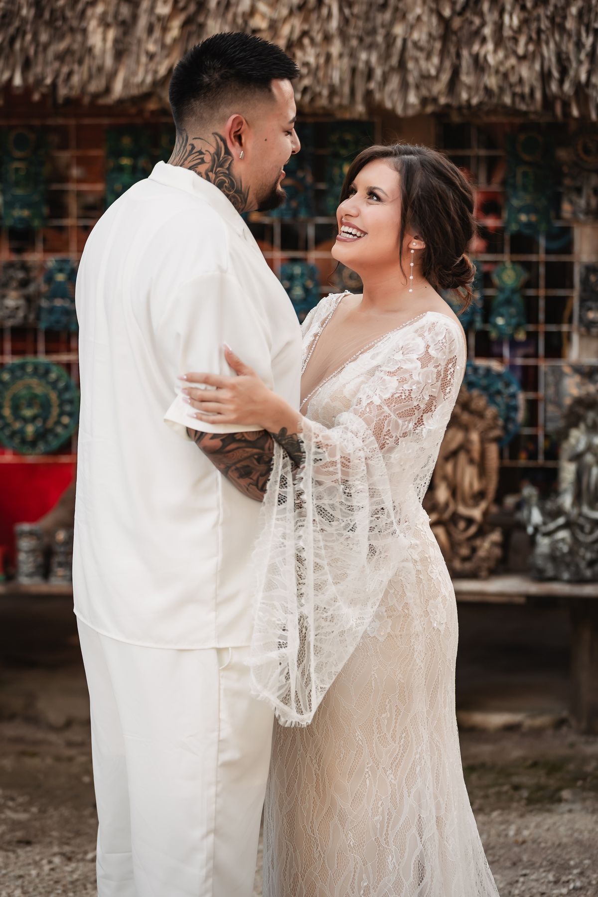 A tender moment between newlyweds in white attire against a traditional thatched building backdrop.