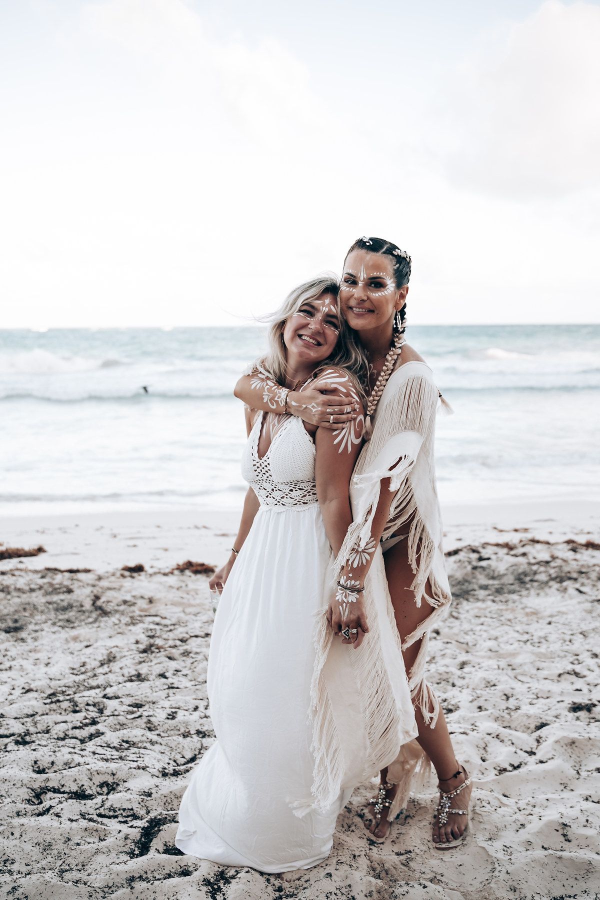 Two people in flowing white dresses share a joyful moment on a beach.