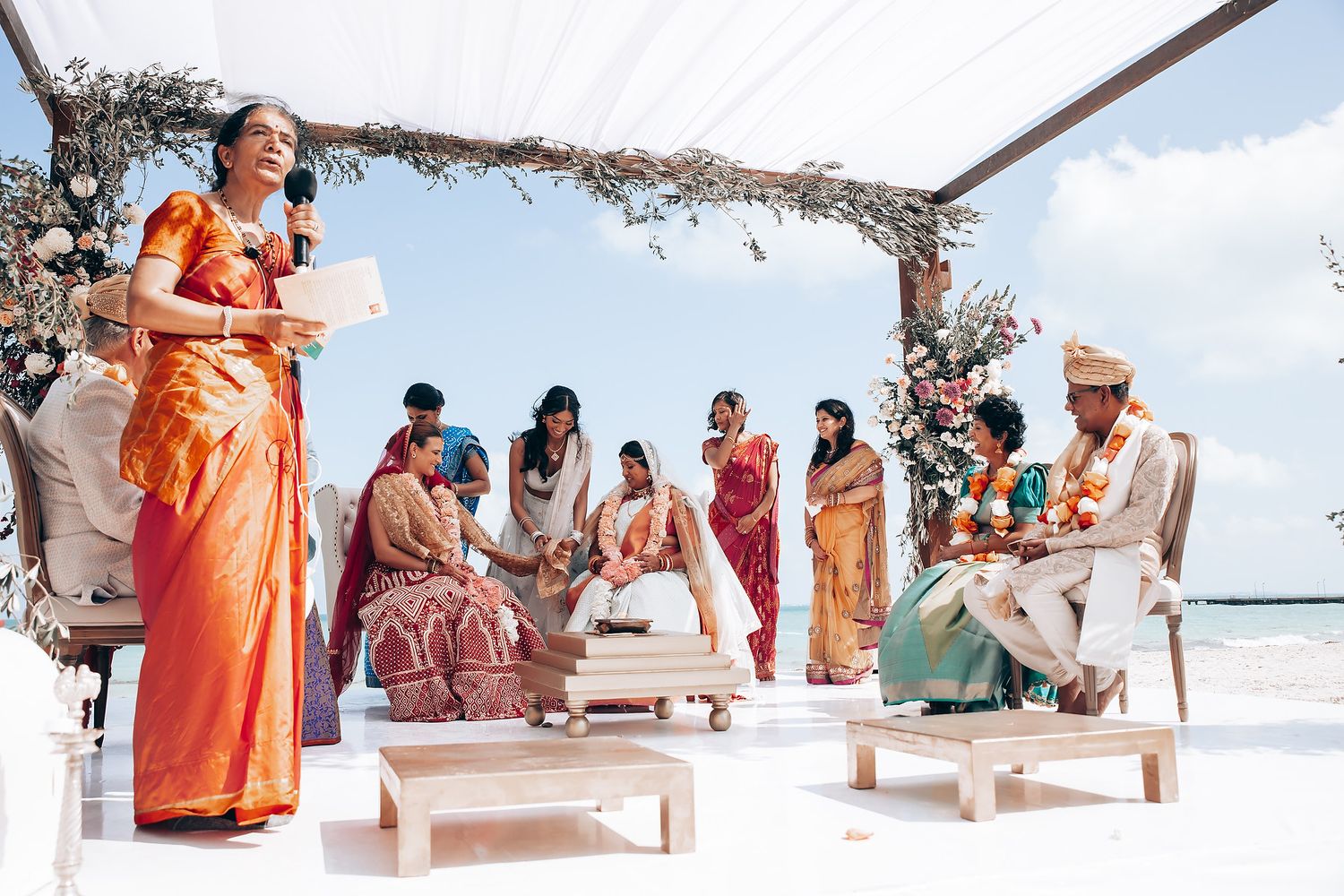 Beachfront wedding celebration with guests gathered under floral-adorned white canopy.