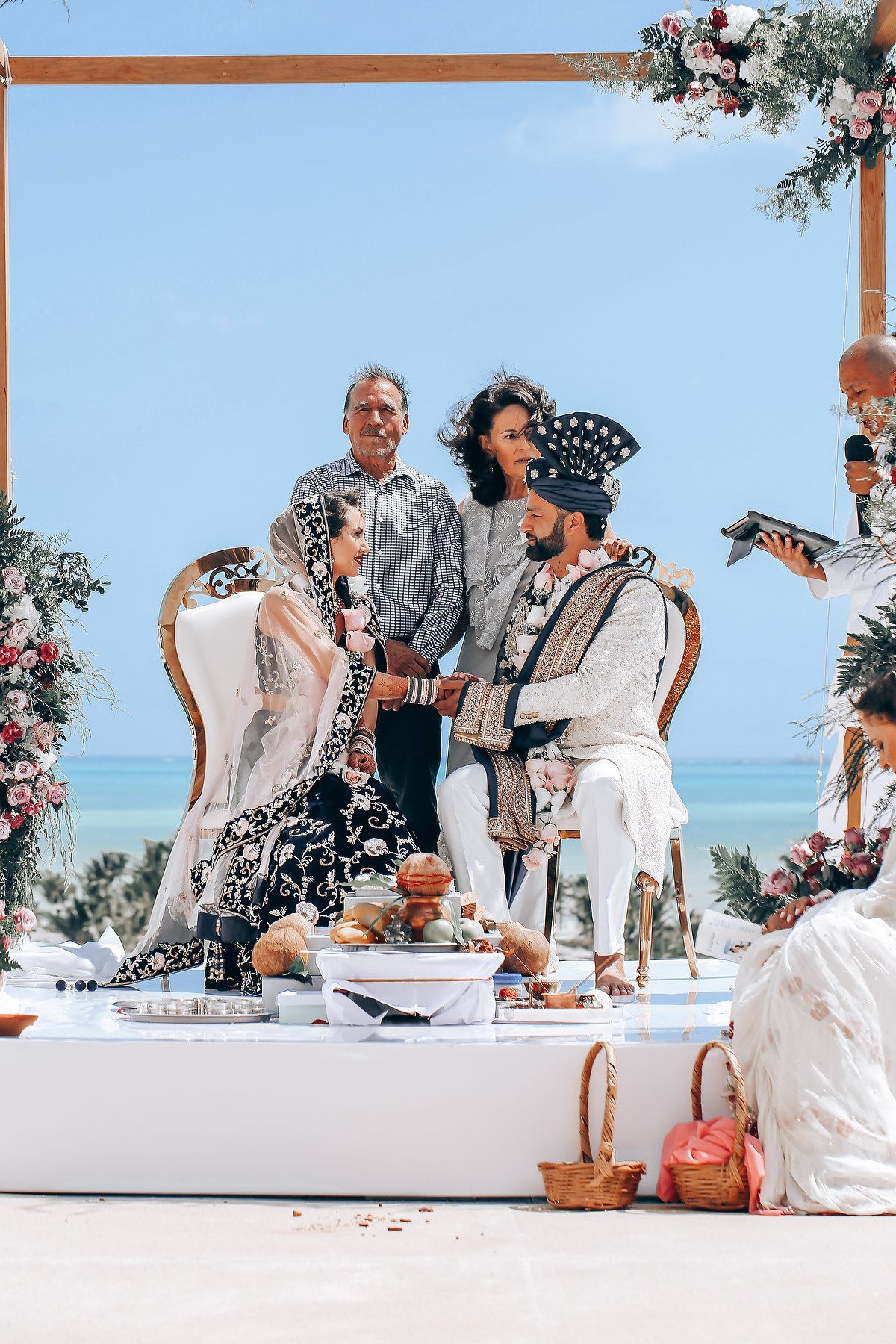 A traditional wedding ceremony takes place under a floral arch overlooking the ocean with guests seated at tables.