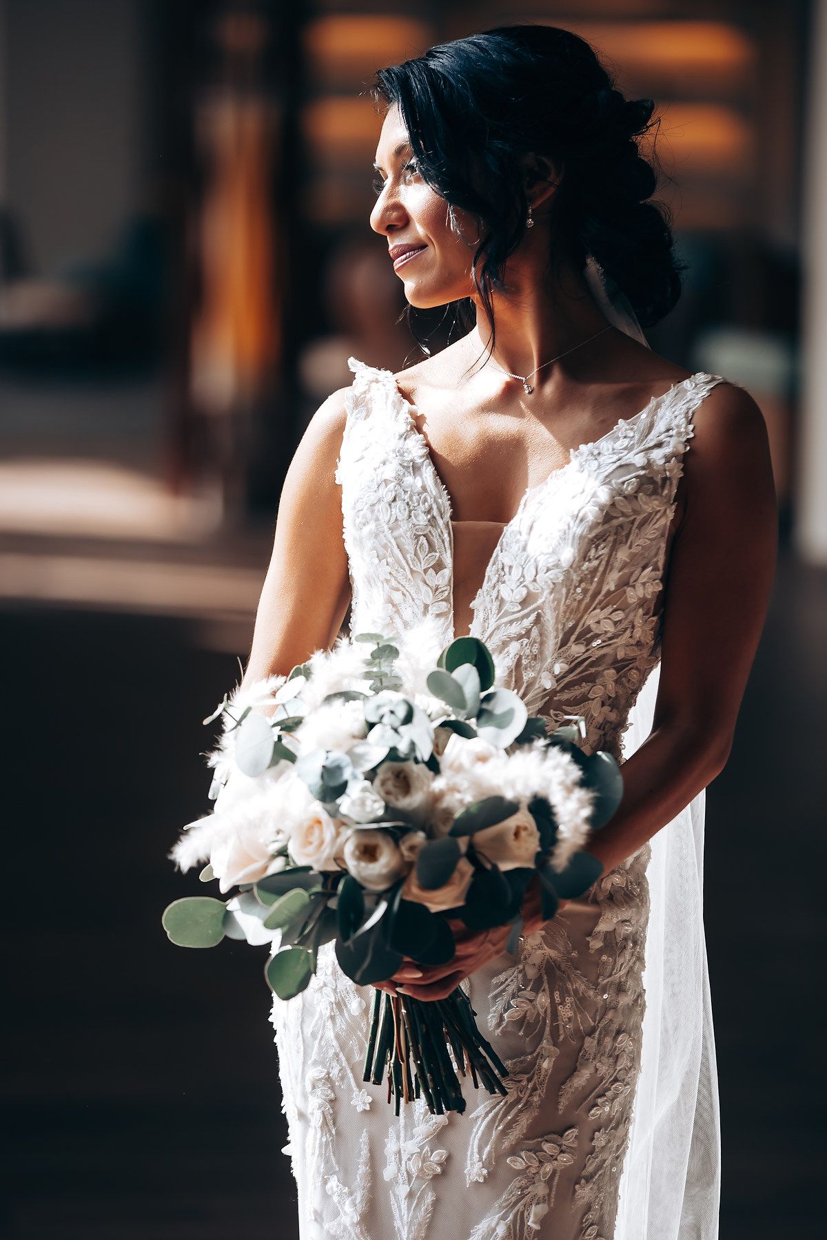 A bride in a lace wedding dress holds a white and green floral bouquet while smiling in soft indoor lighting.