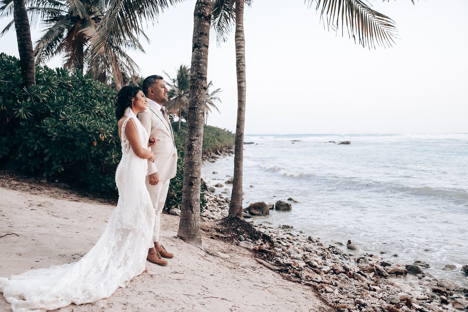 Wedding couple poses along rocky coastline with palm trees and white sand beach.