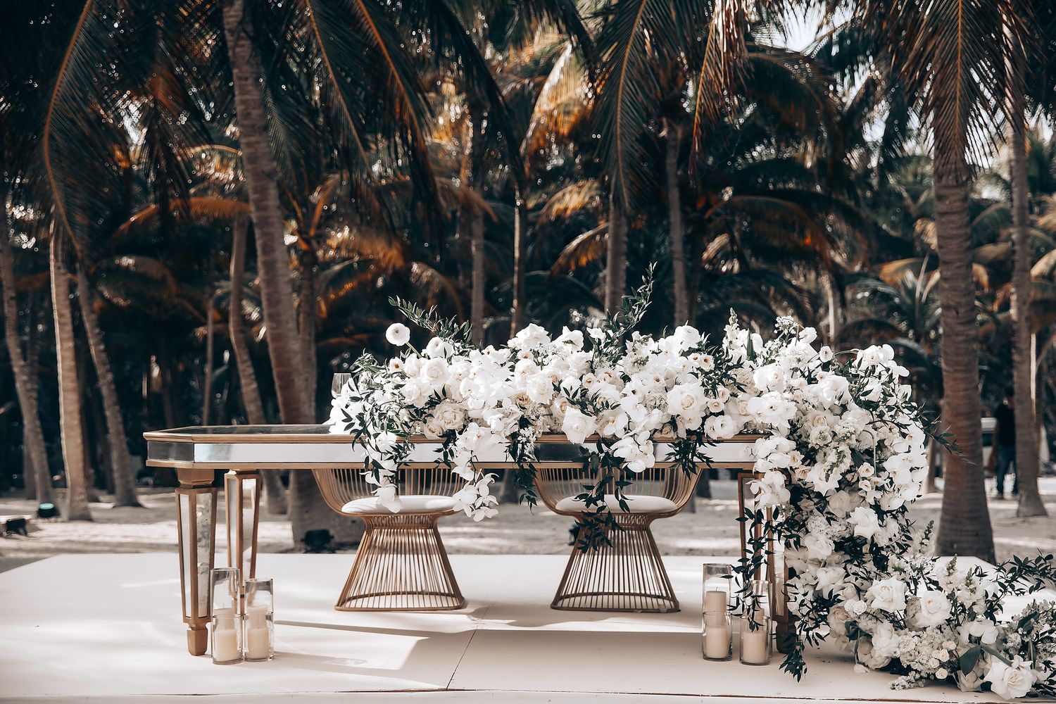 Elegant outdoor wedding reception setup with white floral arrangements and palm trees.