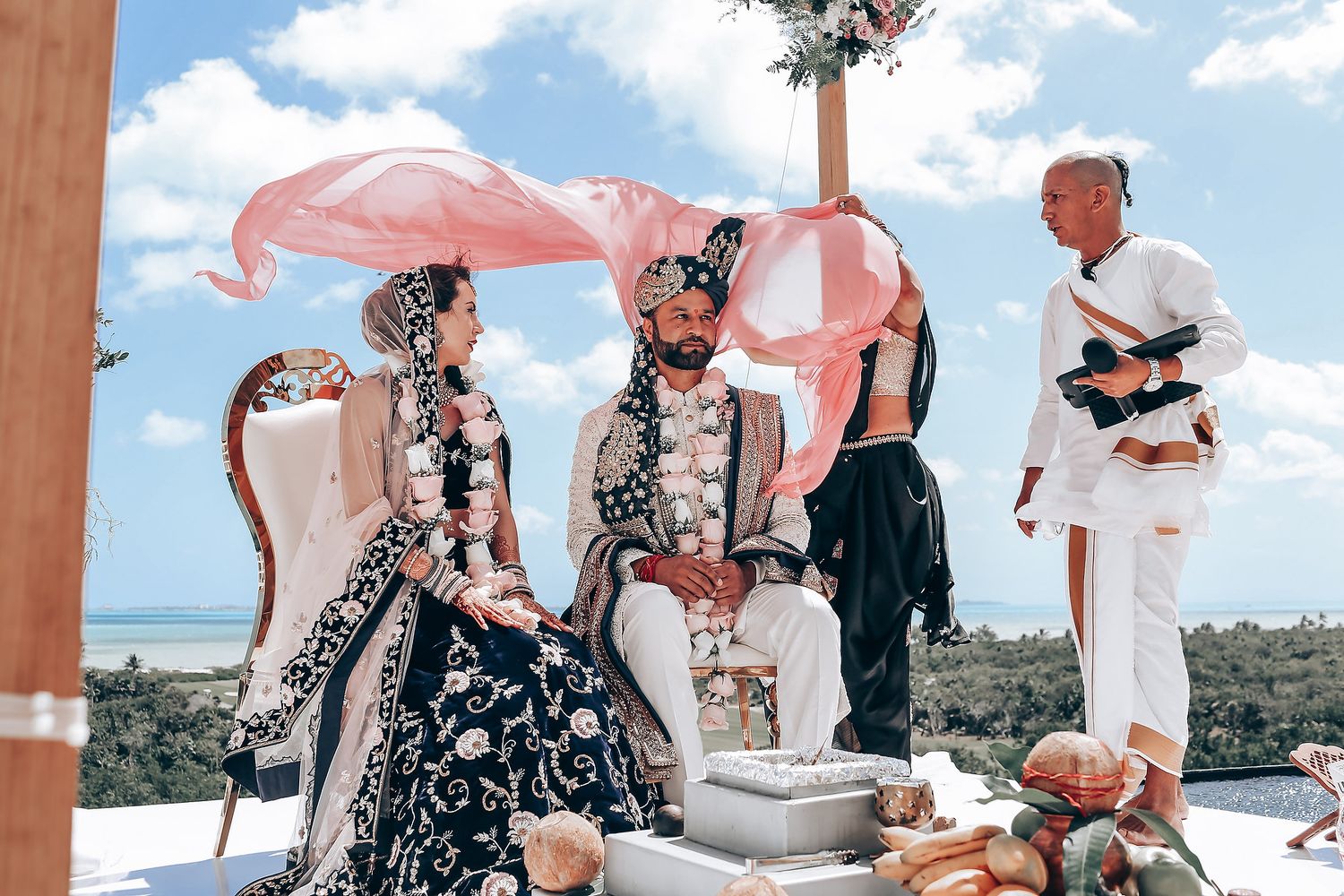 Wedding guests enjoy an outdoor reception with pink parasols against a scenic coastal backdrop.