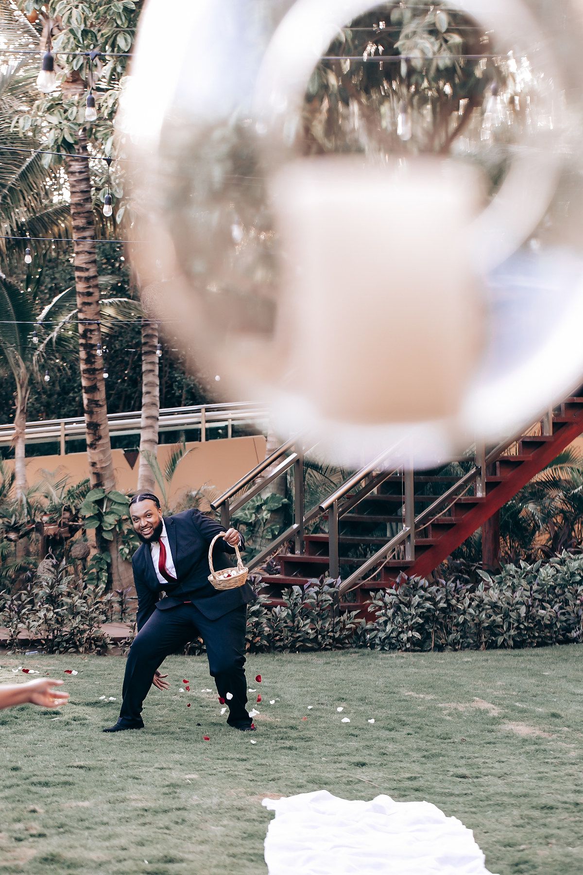 Two people in formal attire playfully pose together in a garden setting with blurred bokeh effects in the foreground.