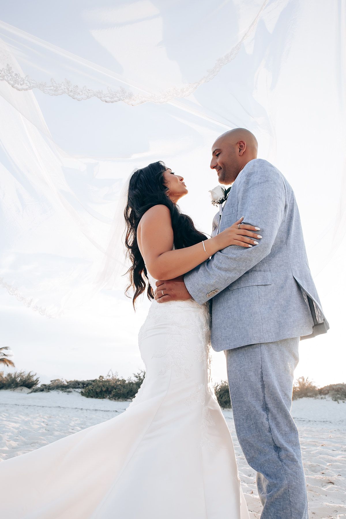 Couple embracing during beach wedding photoshoot against bright sky.