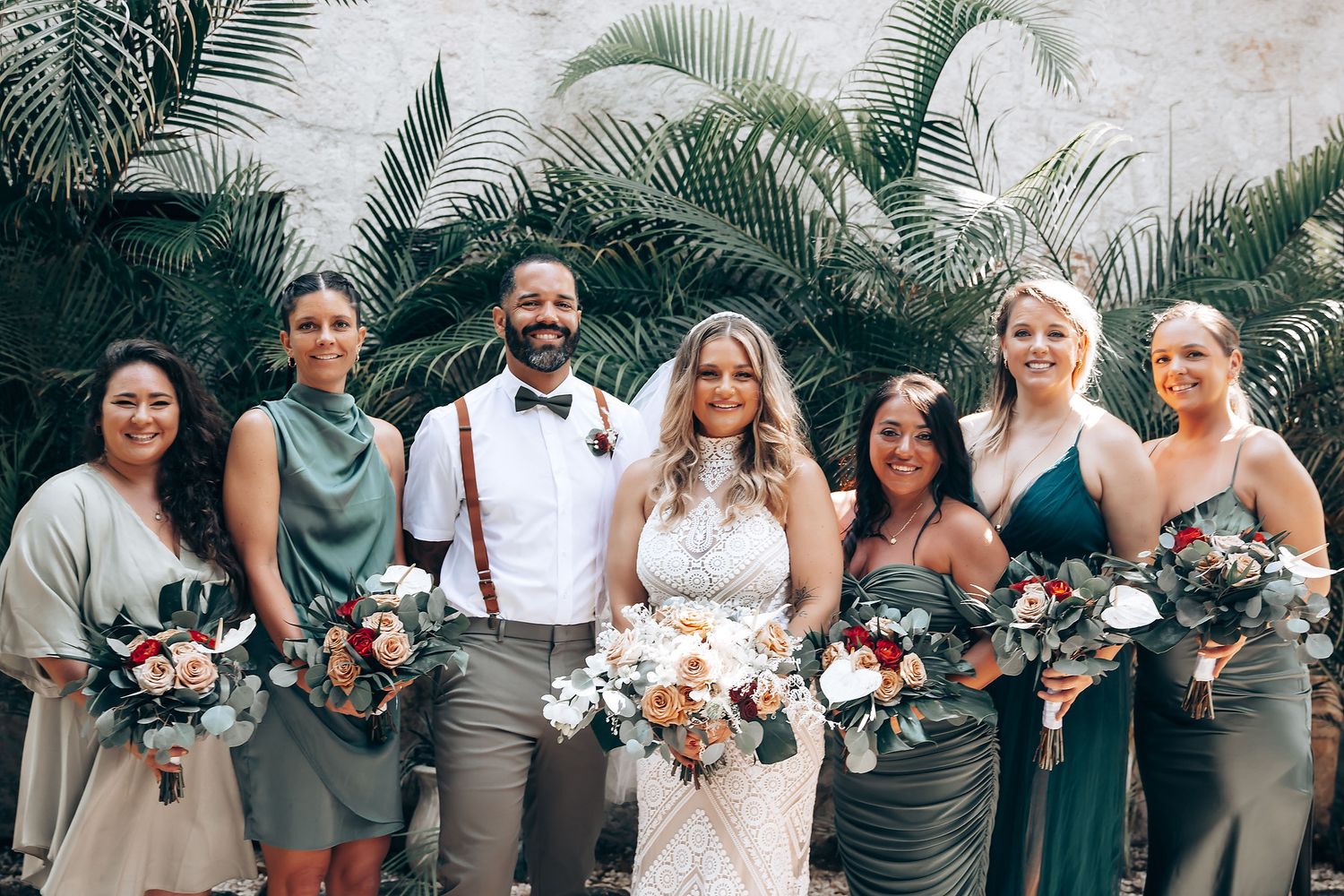 Wedding party poses outdoors with bridesmaids in grey dresses and groomsmen in grey suits holding floral bouquets.