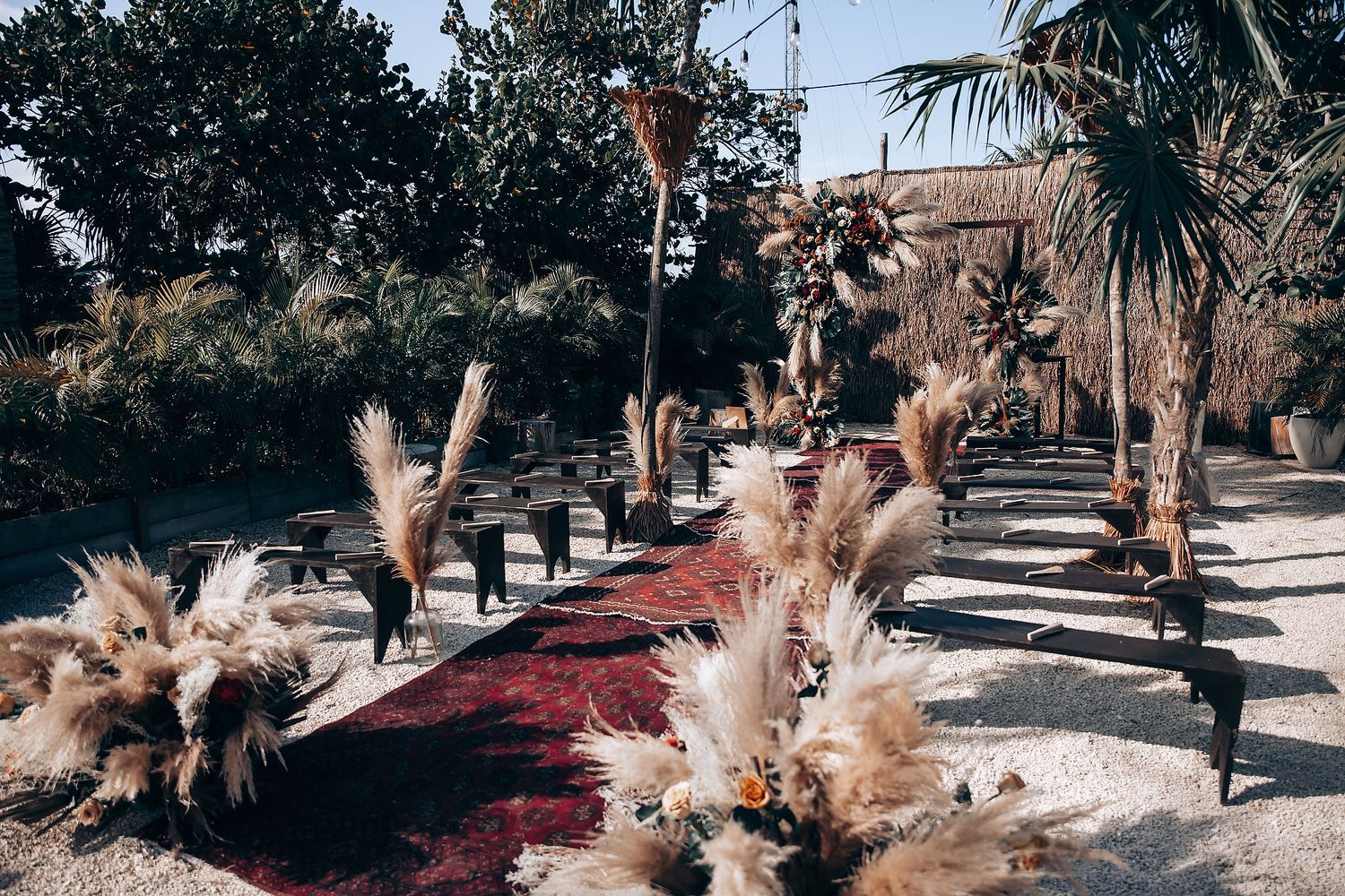 Outdoor wedding ceremony setup with burgundy aisle runner and pampas grass decorations under palm trees.