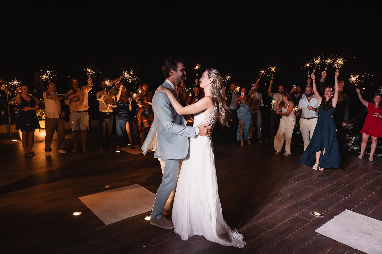 Newlyweds dance together on wooden deck while guests hold sparklers in celebration.