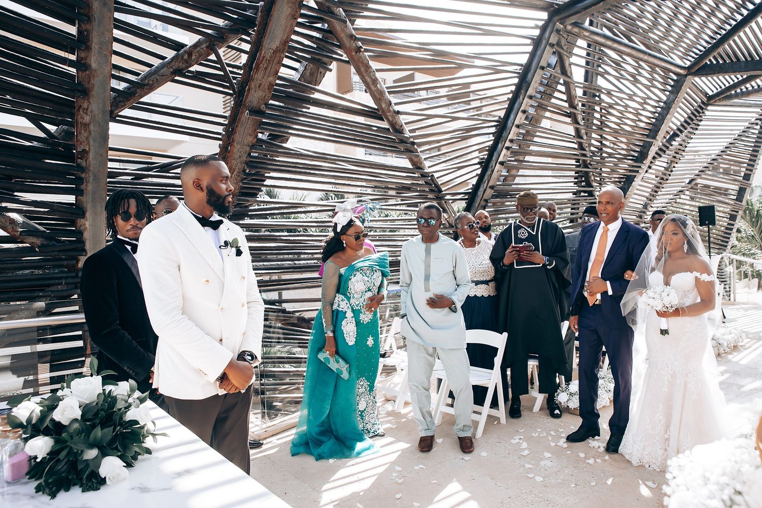 Wedding ceremony guests seated under a slatted pergola roof creating dramatic shadows.