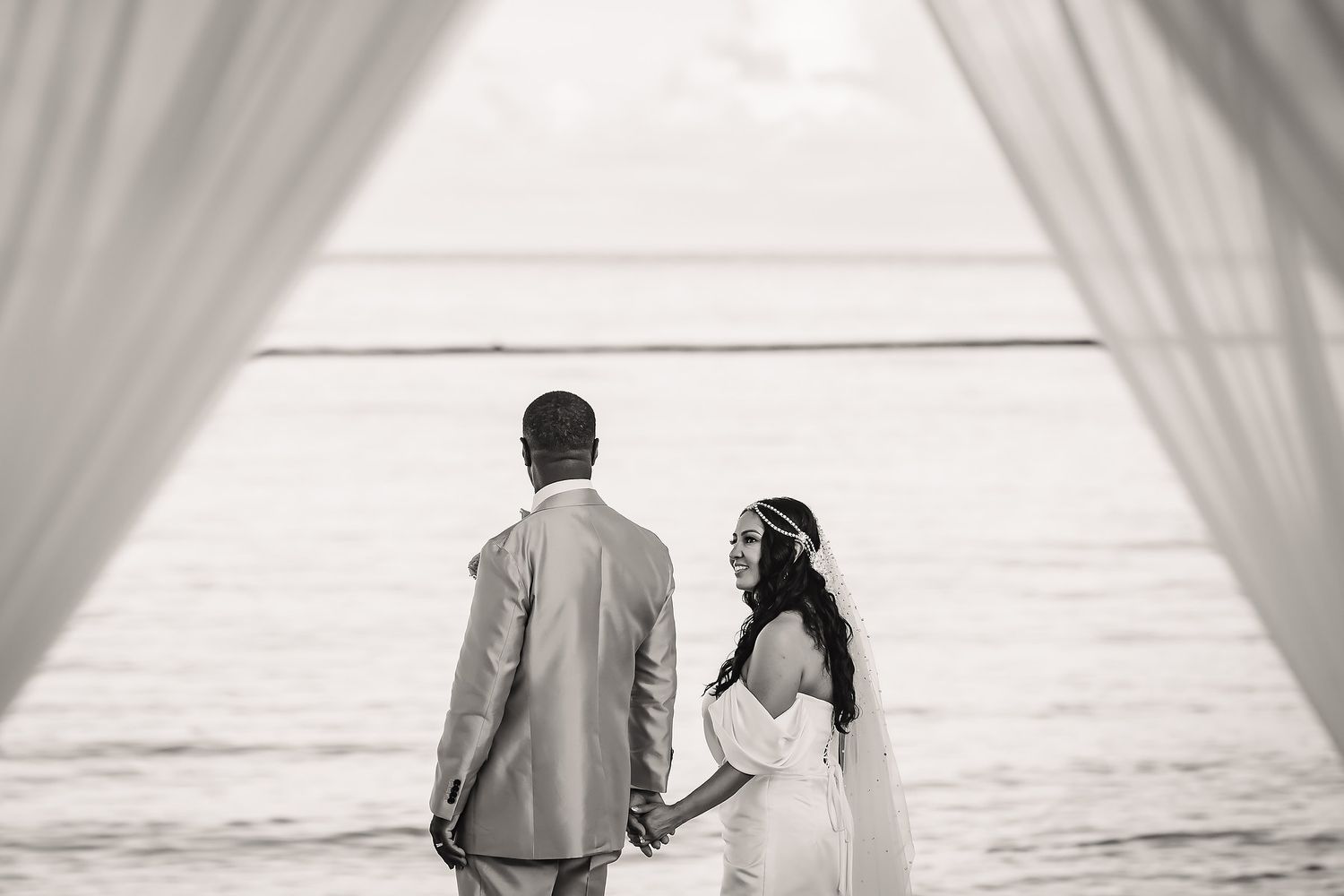 Black and white photo of a couple looking at ocean through white curtains.