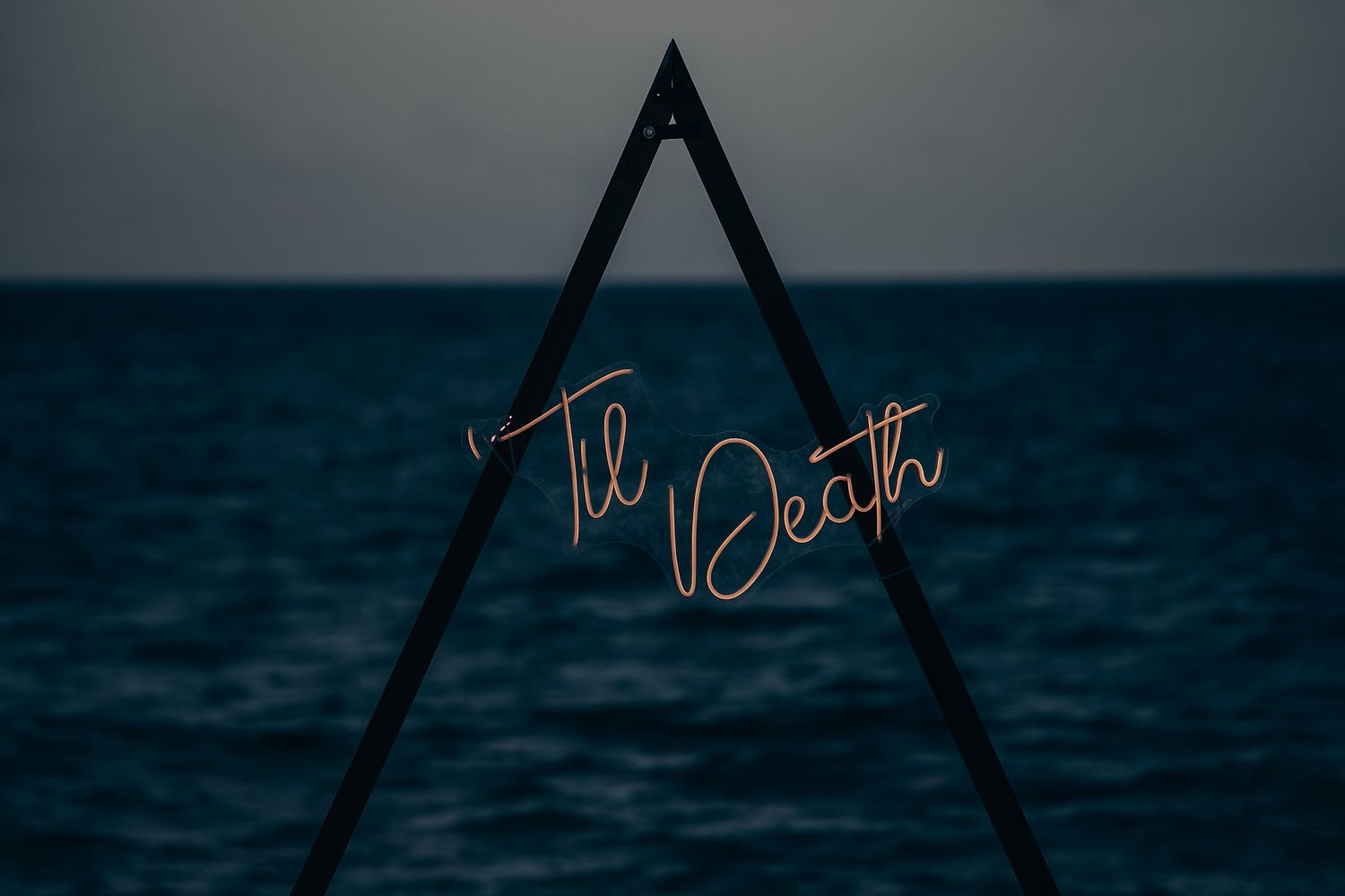Silhouetted triangular neon sign glowing against a dark ocean horizon at dusk.
