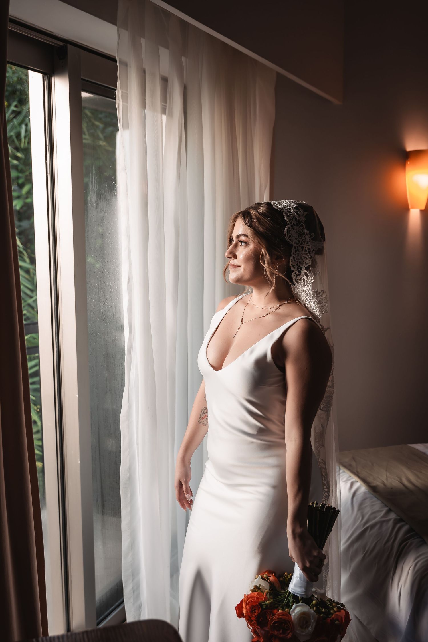 Bride in sleeveless white dress stands near window with dramatic lighting and floral bouquet.