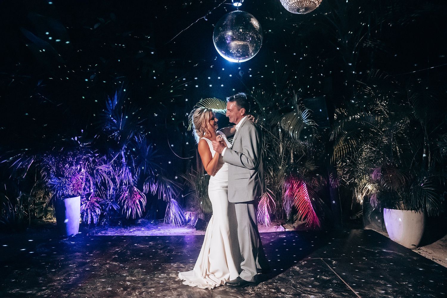 A couple dancing under disco balls with dramatic purple lighting and tropical decor.