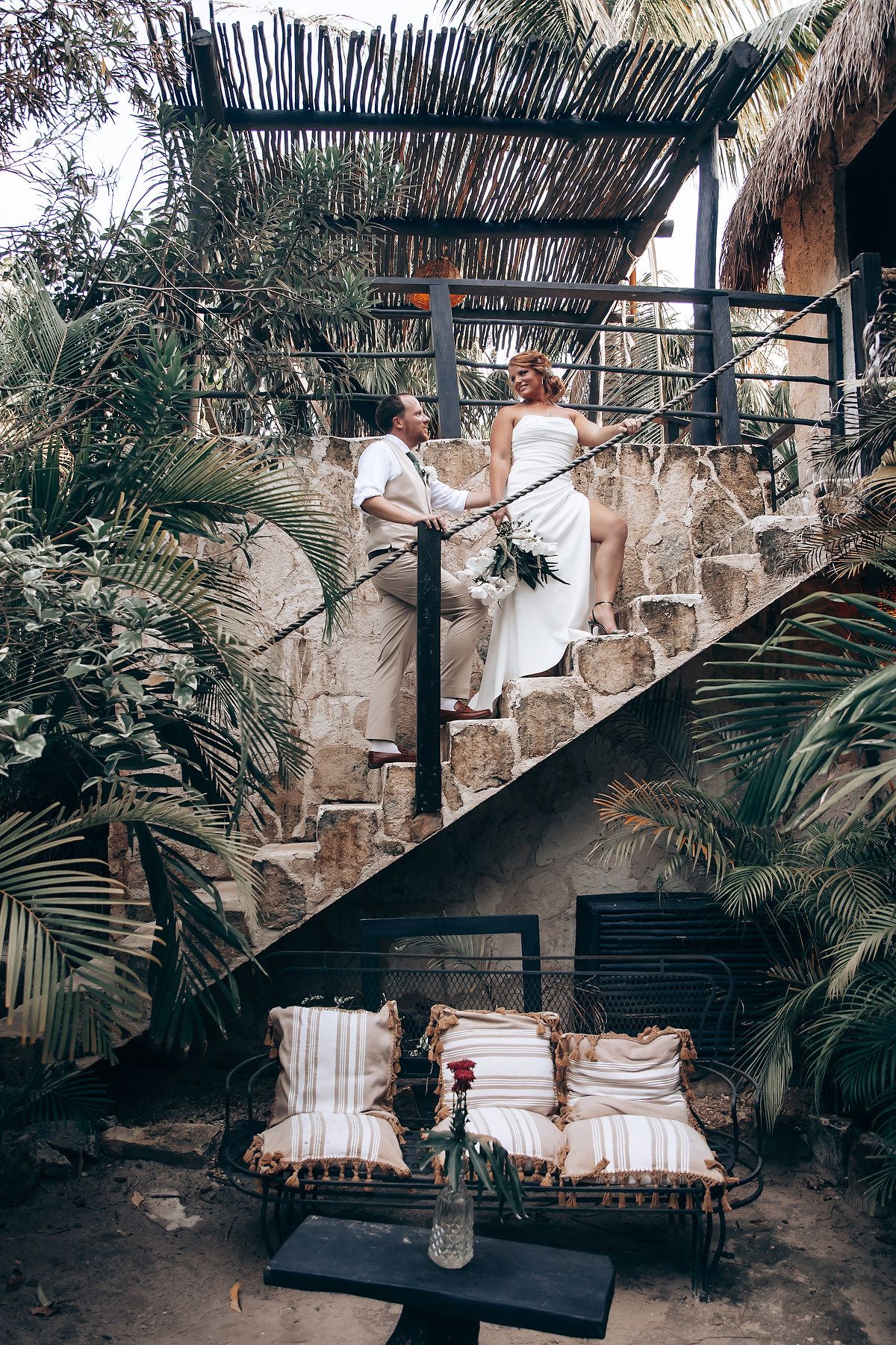 Wedding dress displayed on stone steps surrounded by tropical plants and palm fronds.