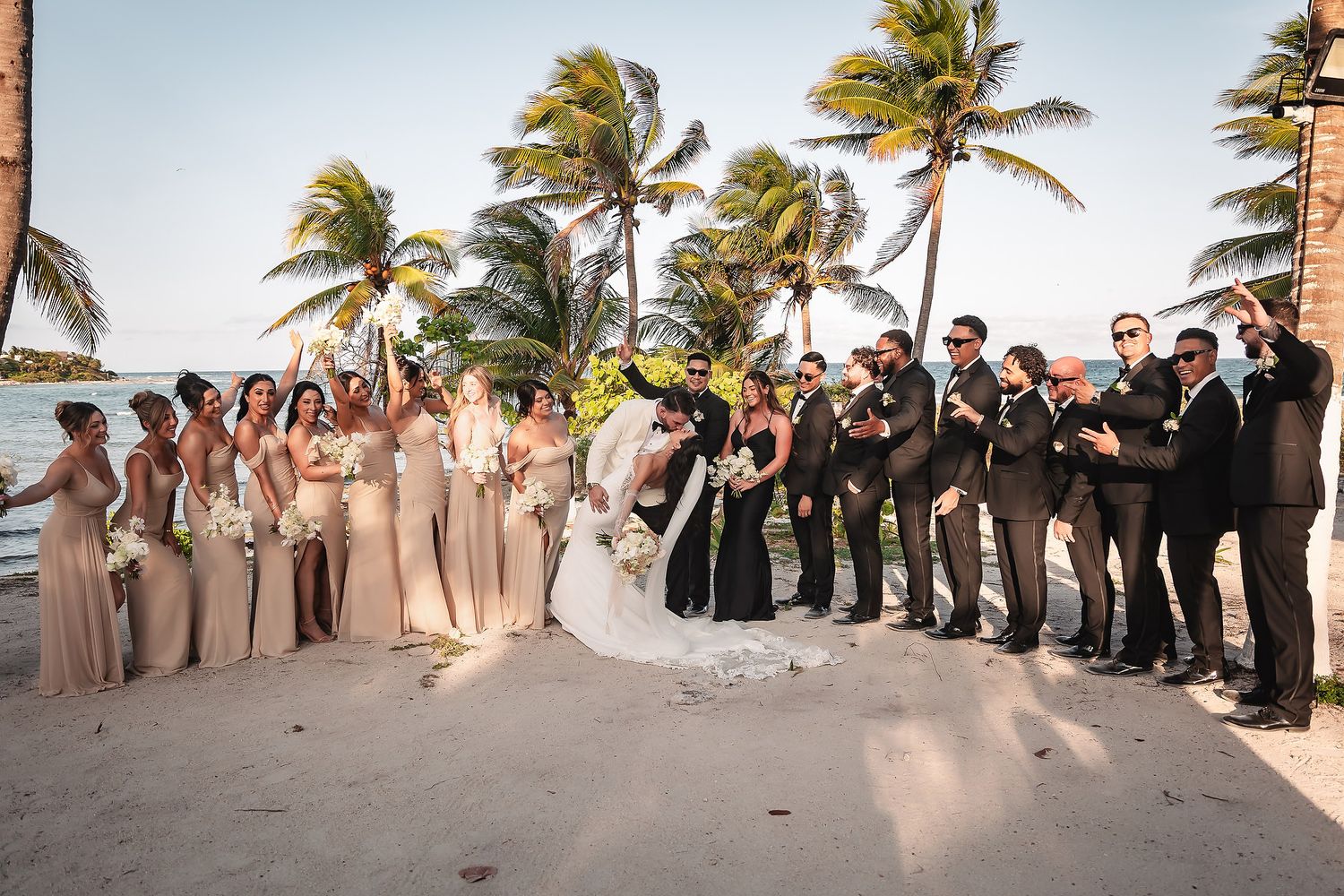 Wedding party poses on beach with palm trees, bridesmaids in beige and groomsmen in black.