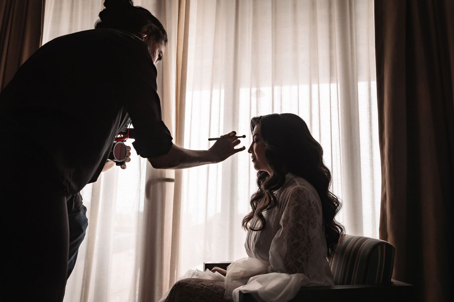 Silhouette of a hairstylist working on long wavy hair in a sunlit room.