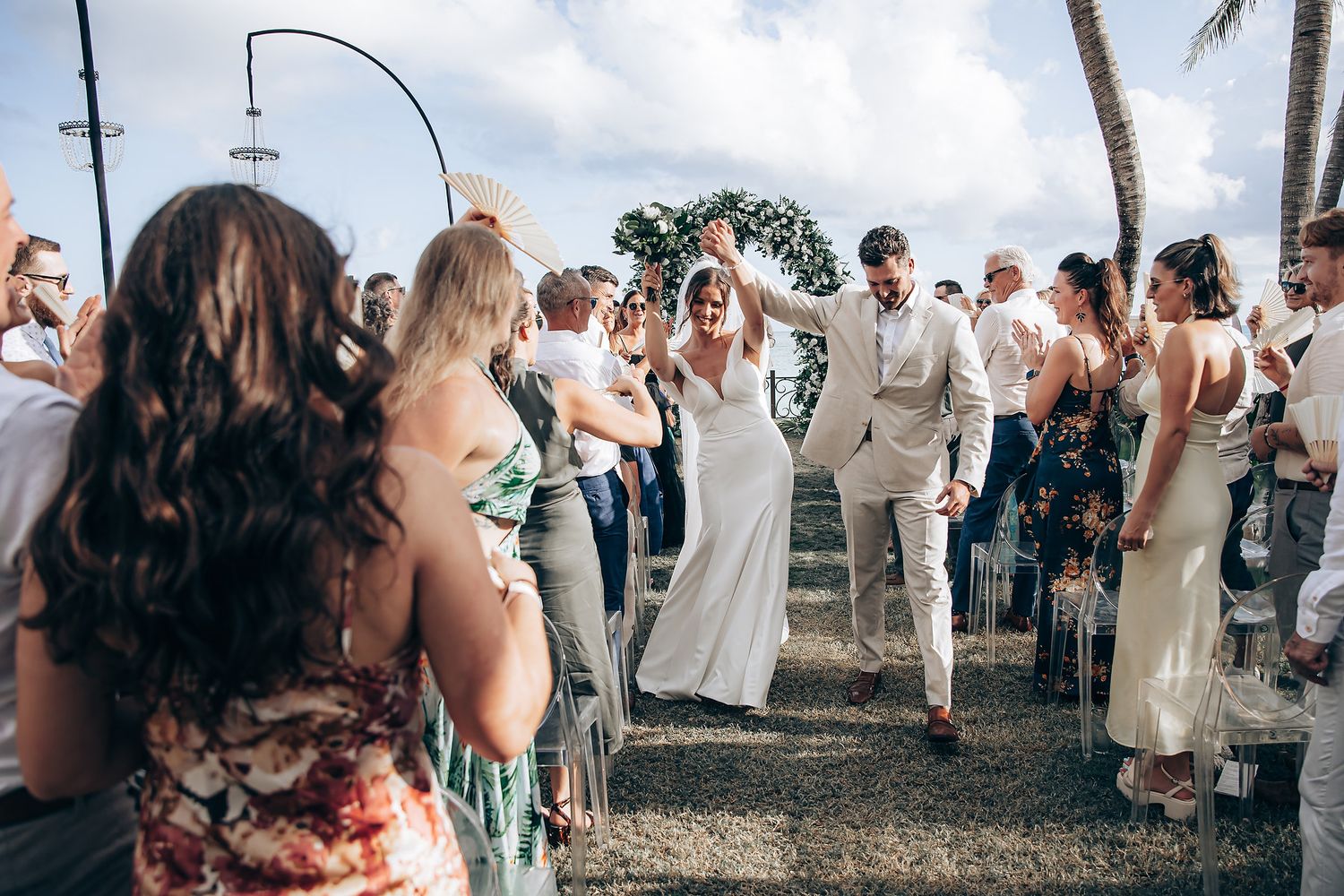 Wedding guests gather for an outdoor celebration with palm trees and ocean views.