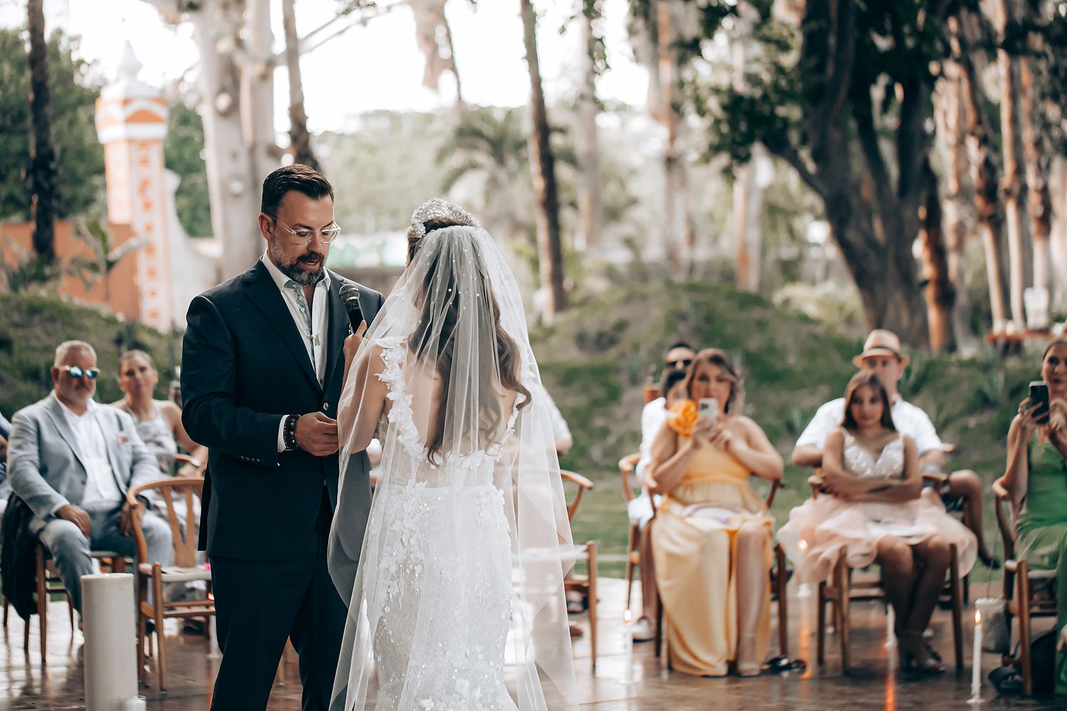 An intimate outdoor wedding ceremony under a canopy of trees with guests seated in white chairs.