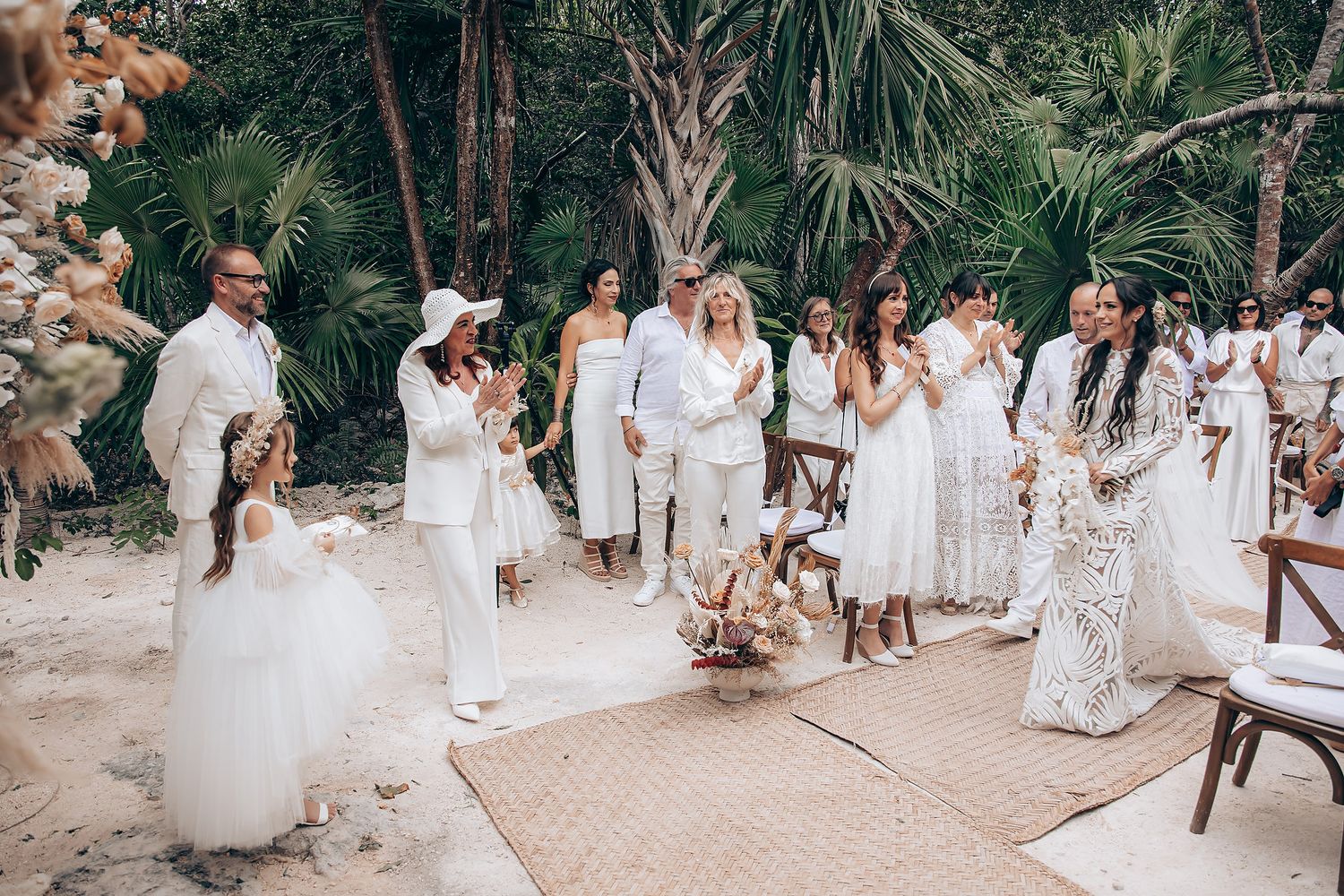 A wedding party dressed in white gathers on a beach path surrounded by tropical palm trees and greenery.