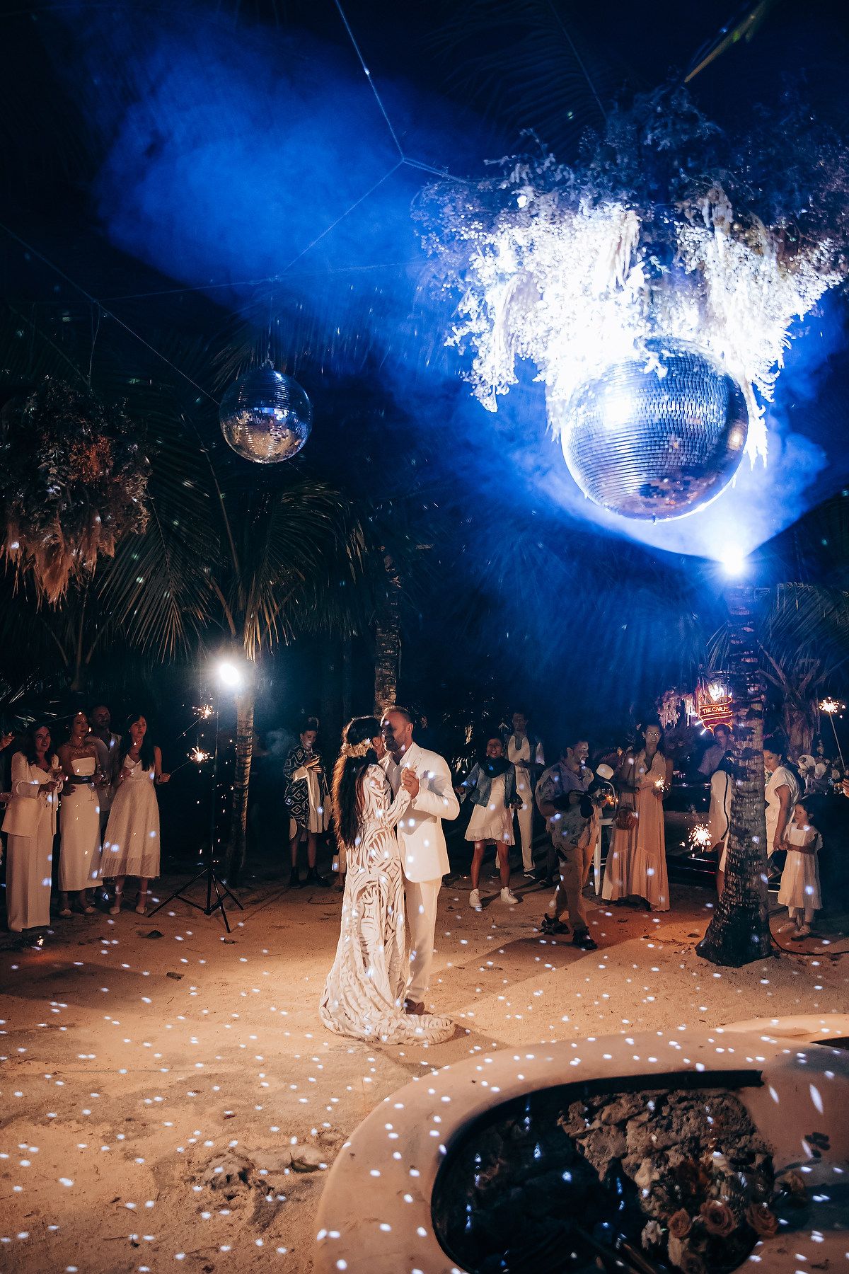 A couple shares their first dance under dramatic blue lighting with flower petals scattered on the ground at an outdoor wedding.