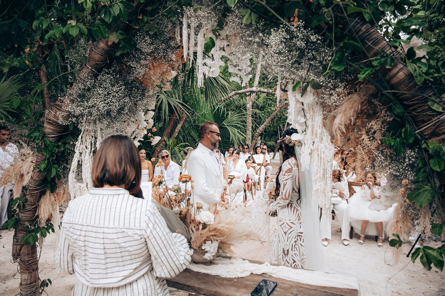 A couple in white outfits walks through a lush garden pathway during an outdoor wedding ceremony.