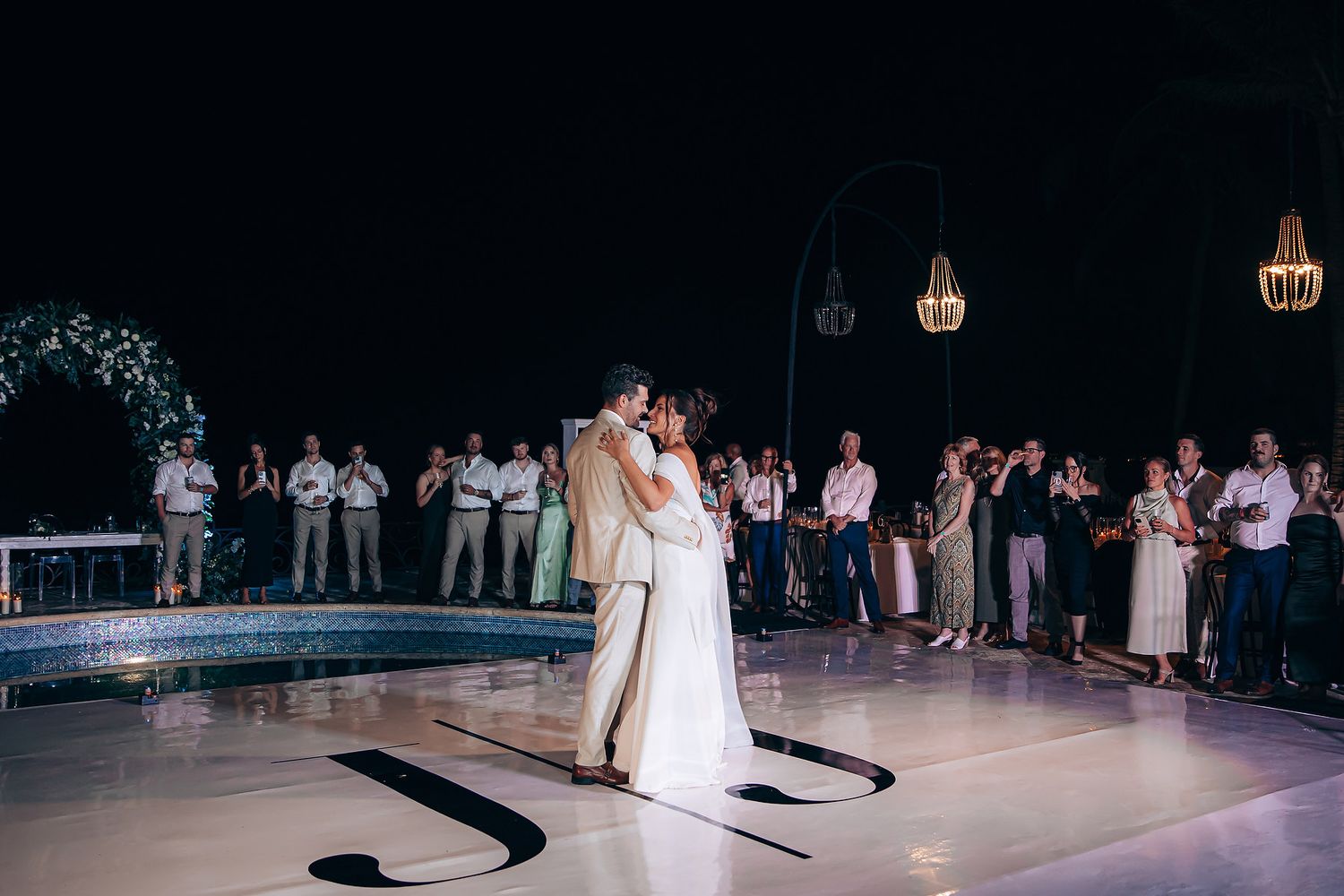 A couple shares their first dance at an evening wedding reception on a white dance floor surrounded by guests and lanterns.