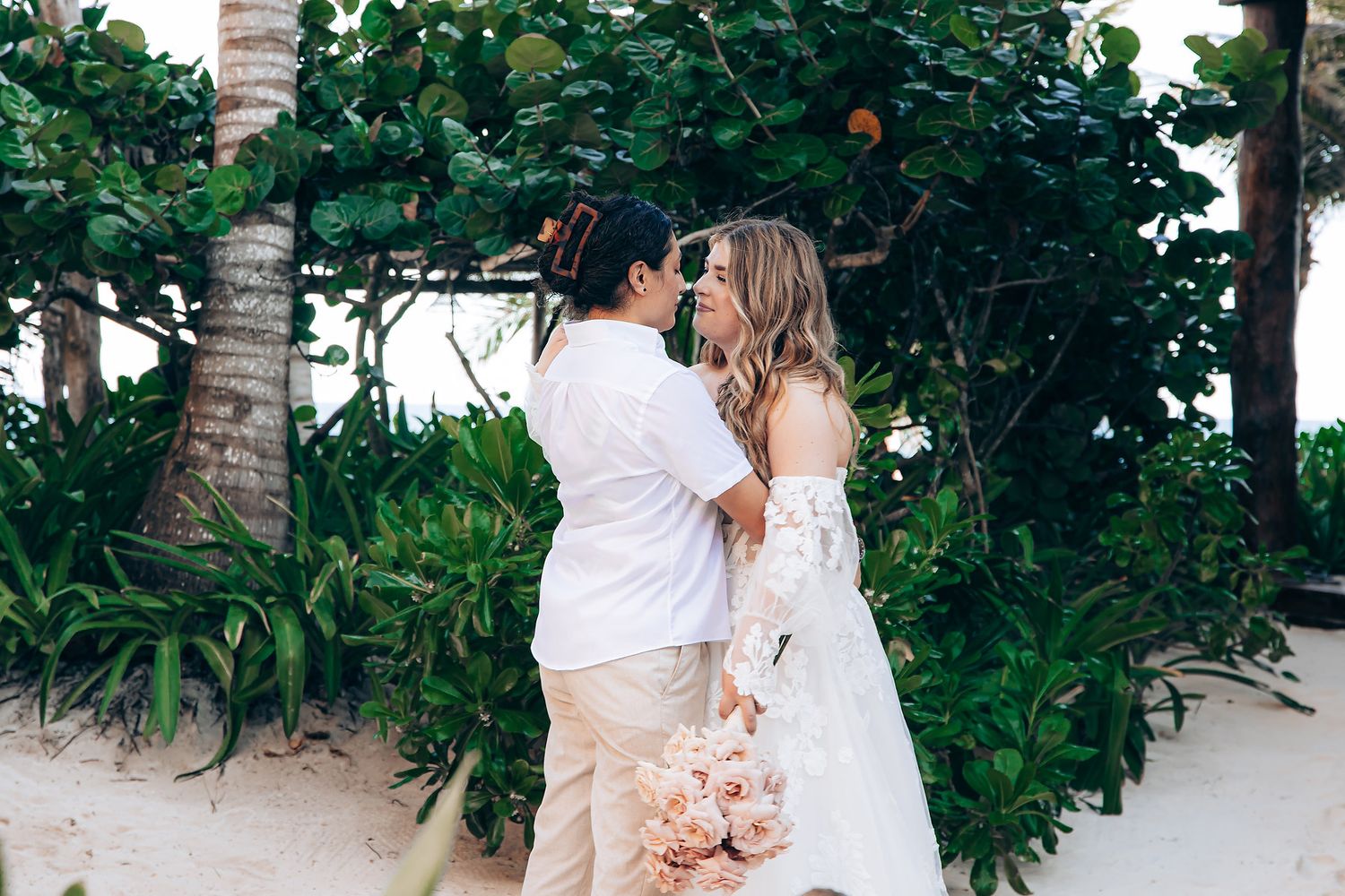 Romantic beach wedding photoshoot with couple embracing among tropical palm trees and lush greenery.