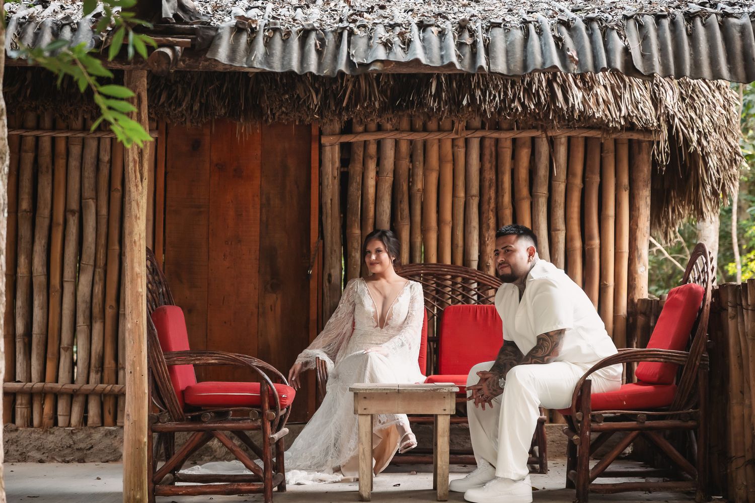 A couple seated in red chairs under a rustic thatched roof structure during their wedding.