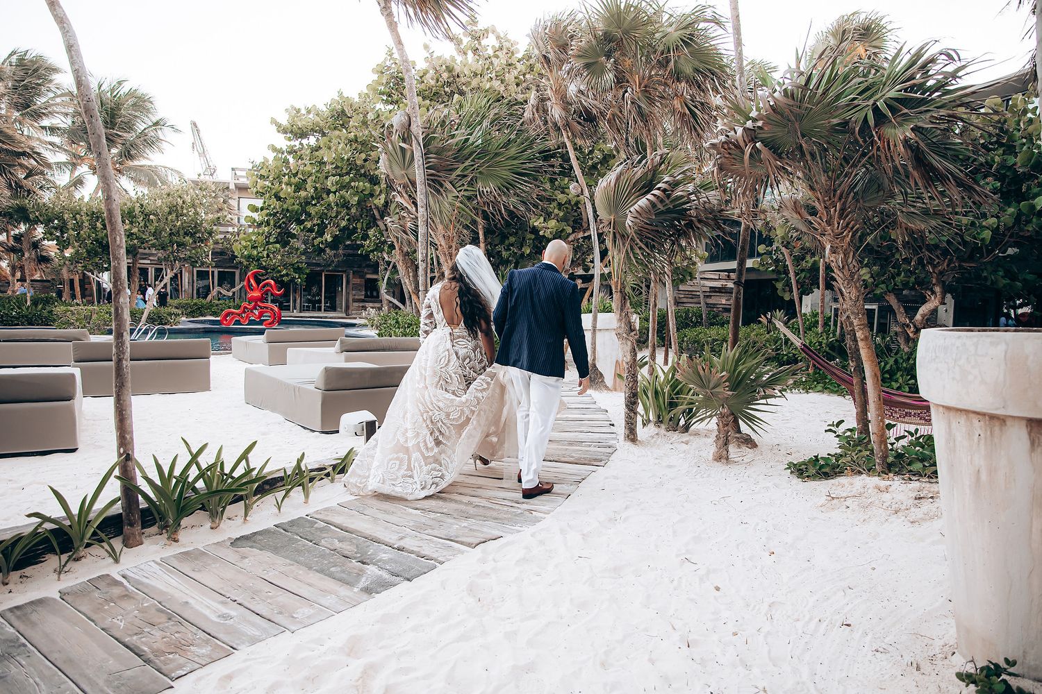 Palm trees line a white sandy pathway along a tropical beach resort with wooden deck seating areas.