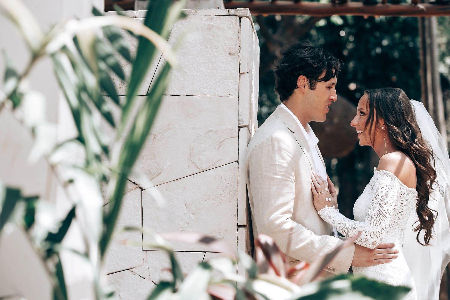 A romantic couple in white attire share an intimate moment against a light colored wall with greenery.