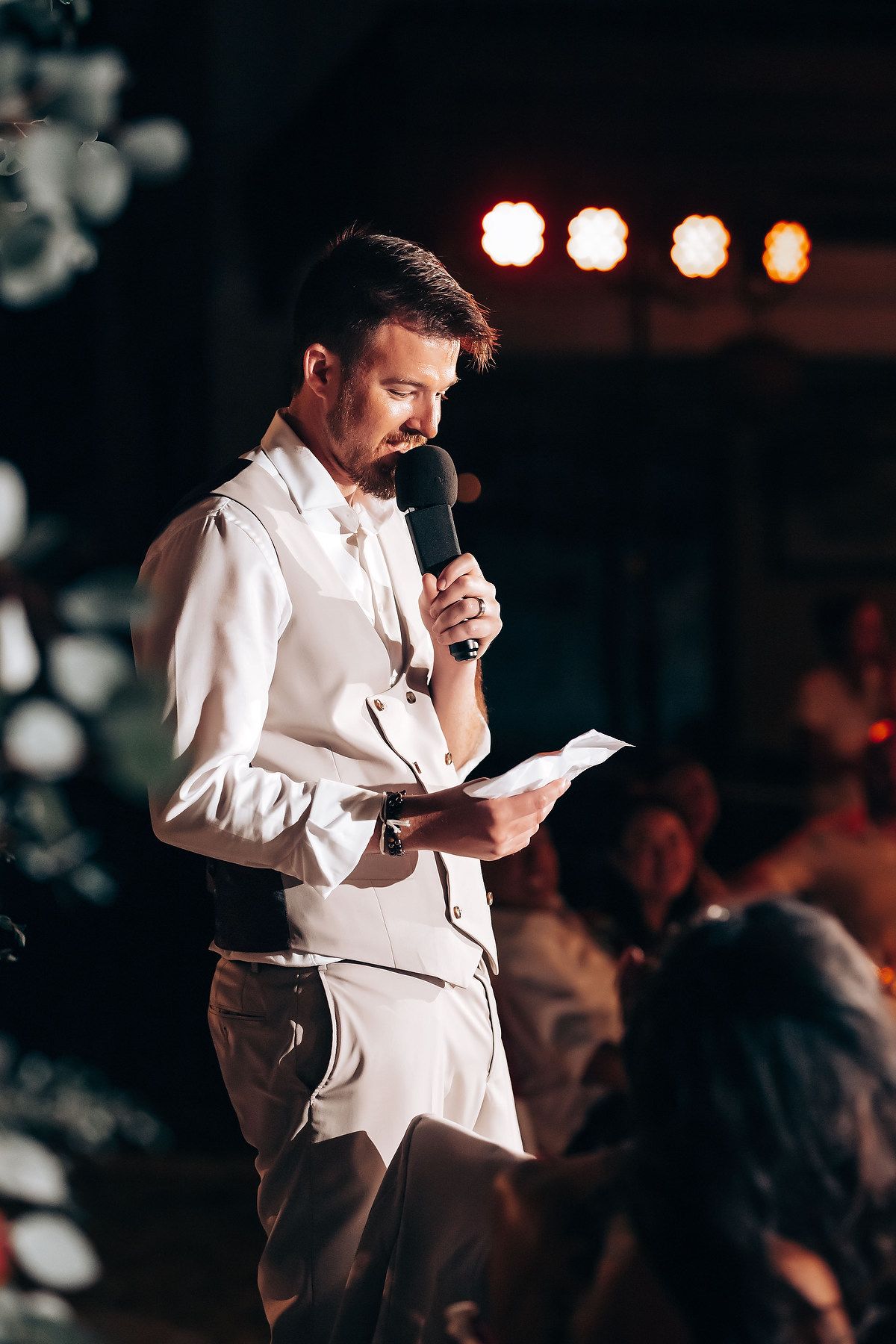 Person in white suit reading from paper during evening wedding reception.
