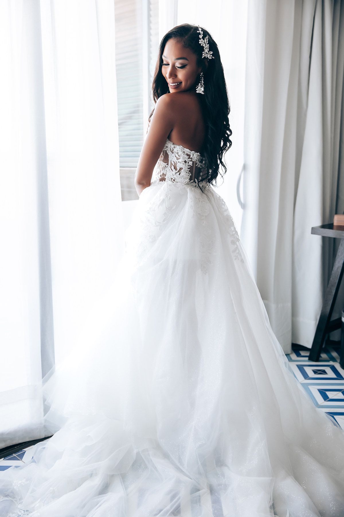 Bride in flowing white wedding gown poses elegantly by window with natural light streaming through sheer curtains.