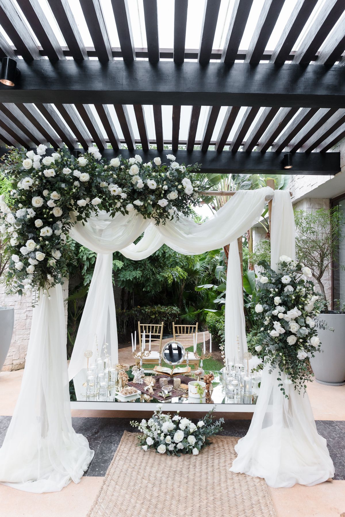 Elegant outdoor wedding arch adorned with white roses, greenery, and flowing drapes over a decorated reception table.